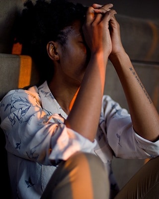 An African American woman sits in a dark room, covering her face and eyes with her hands, as though to manage headache pain.