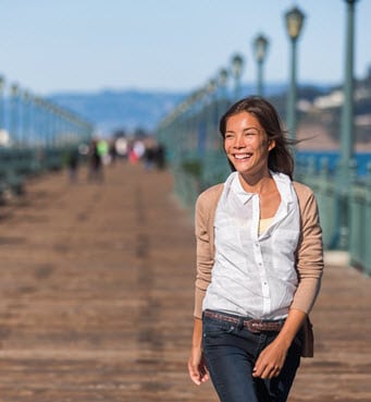Asian American professional woman in jeans and a button down walks along a San Francisco pier on a breezy day.