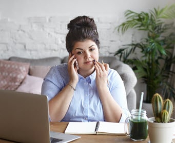 Millennial professional woman talking on her cell phone with a notebook and laptop in front of her. She is a plus sized white woman in a button down and long hair in a bun.
