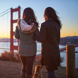 Lesbian Couple stands in front of the Golden Gate Bridge in San Francisco, with their small dog. It is a windy day and they are wearing coats, with their backs turned to the camera.