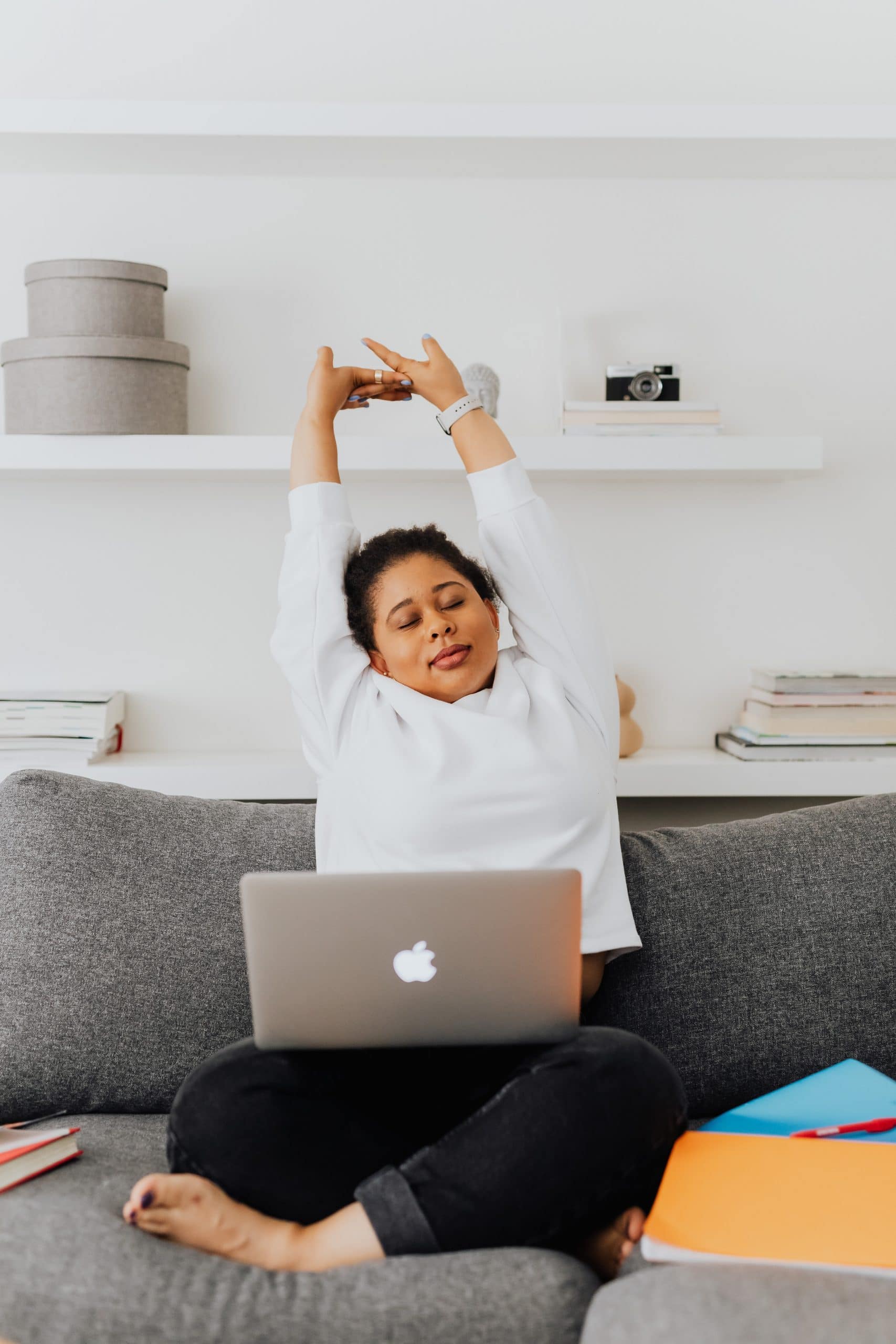 A Filipinx woman working from home with a laptop on the couch. She has a slight smile on her face, eyes closed, and she is stretching overhead, as though she is taking a mindfulness or yoga break during the workday.