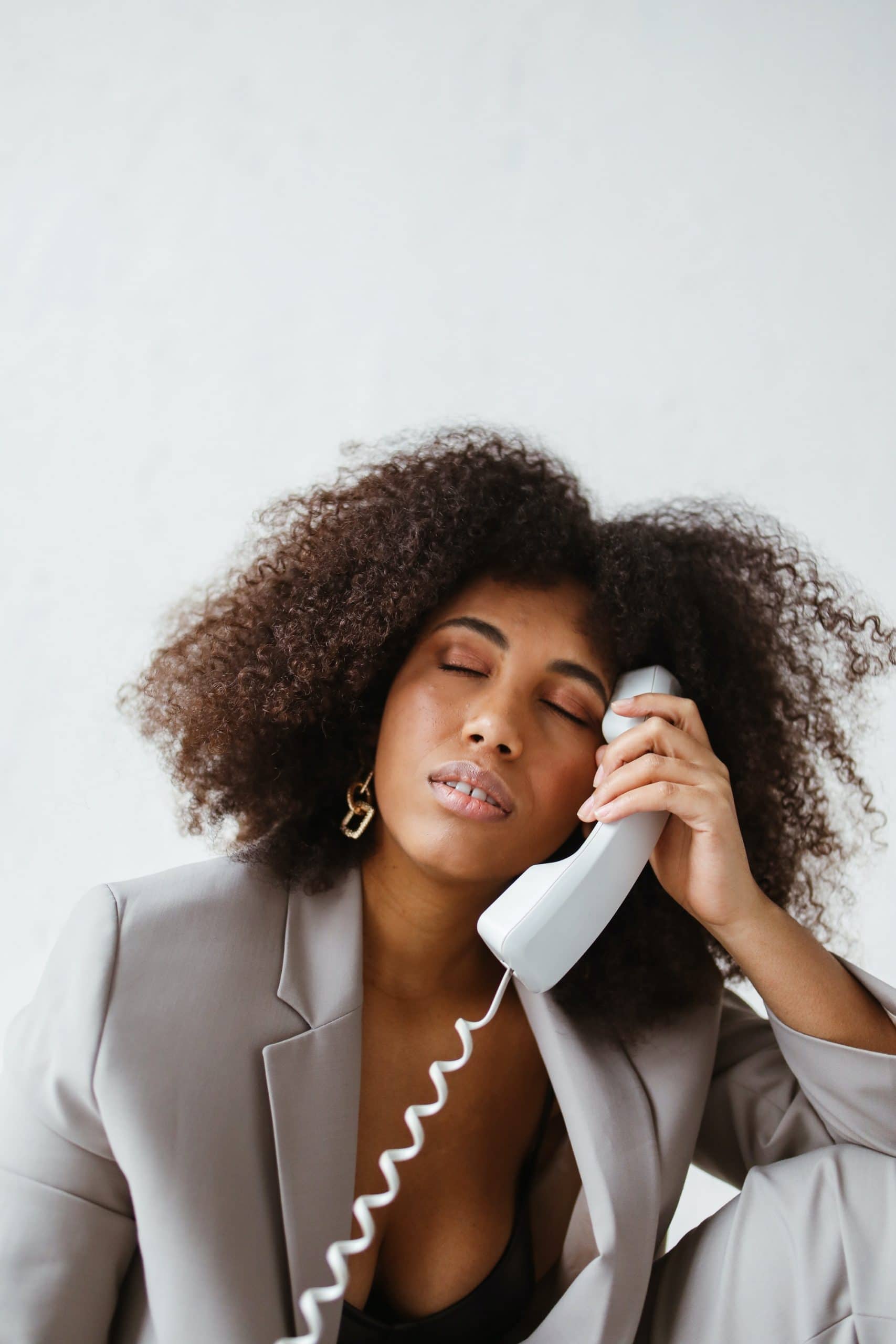Black Millennial professional woman with natural hair in a gray pantsuit, holding a phone receiver. Her eyes area closed and her expression is stressed, exhausted, and burned out.