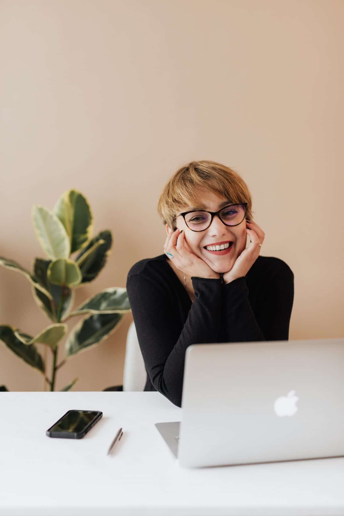 Young short-haired professional woman with glasses sits in front of her laptop. She is holding her head in her hands and is smiling.