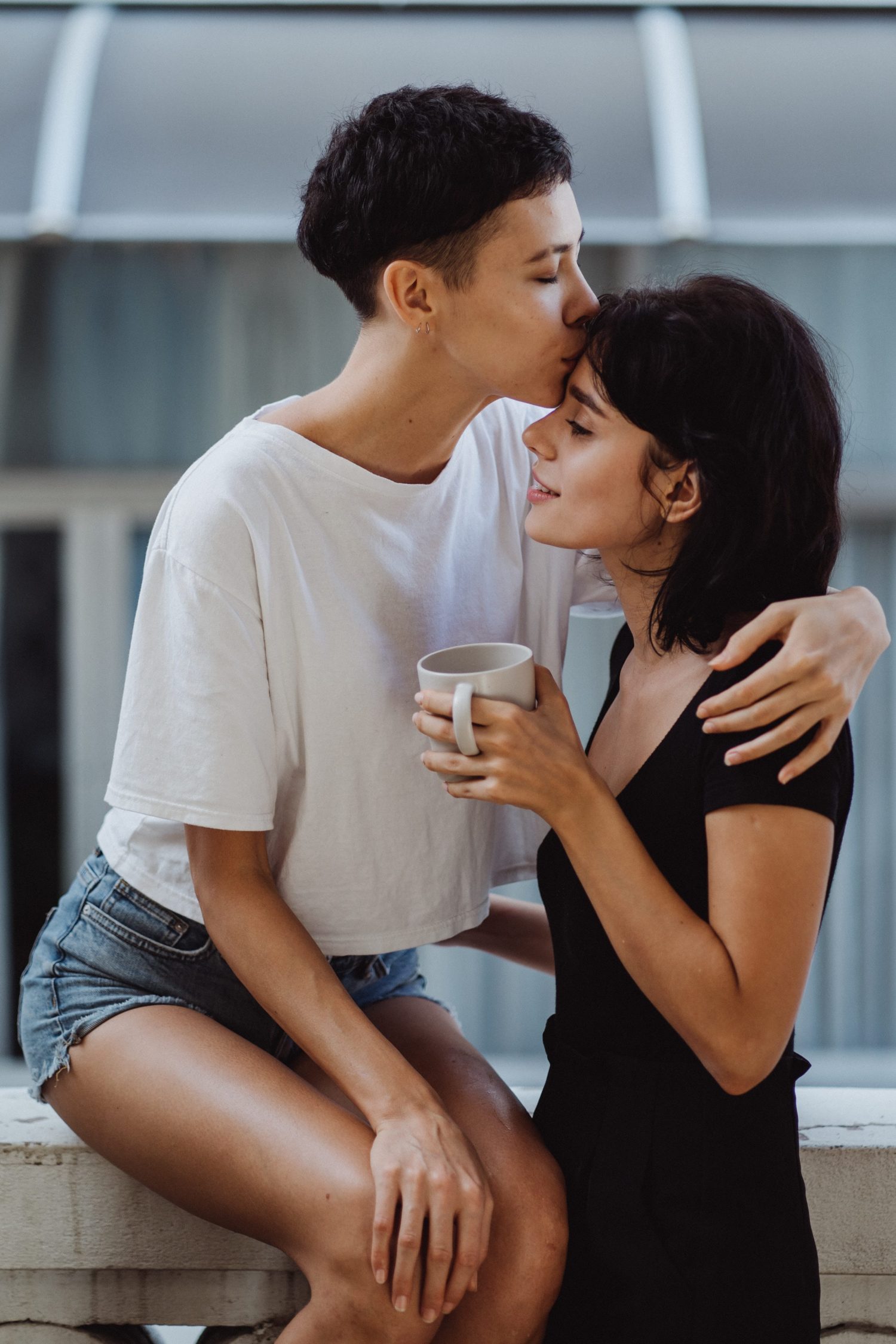Photo of a queer couple. An androgynous person with short hair sits on a ledge, and is kissing the forehead of a femme presenting person in a black dress standing to their right. Both partners have brown skin and dark hair.
