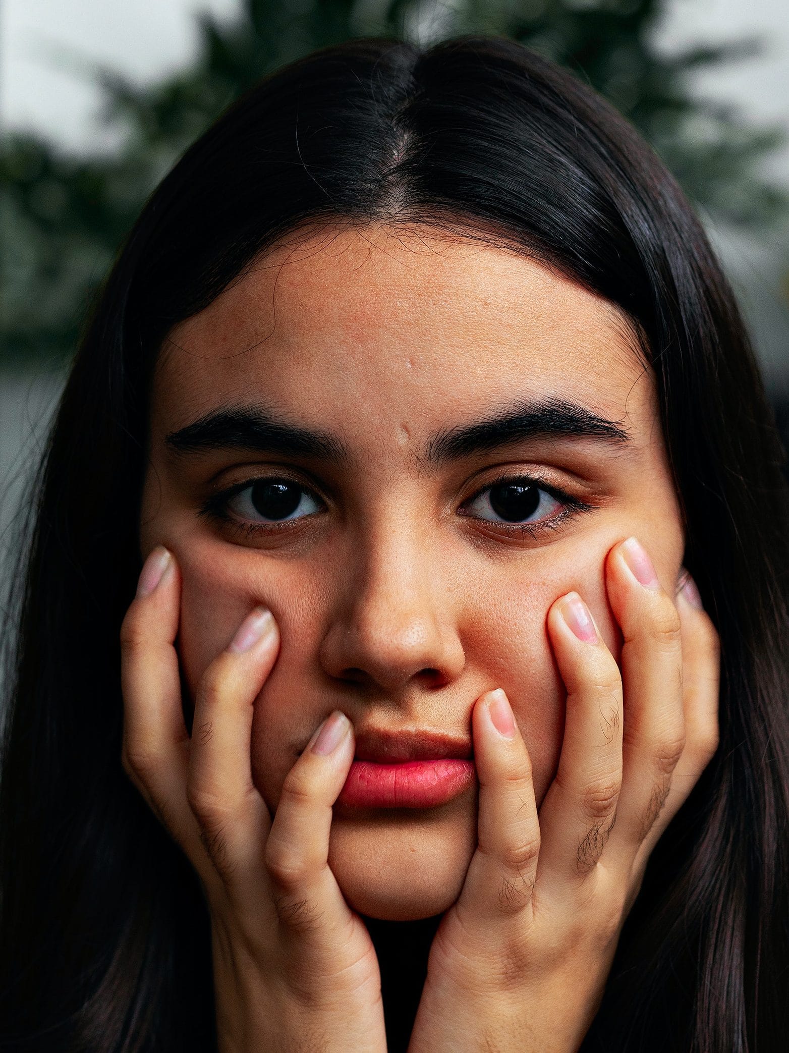 A brown skinned Latinx woman with dark hair is photographed close up. She is holding her face in her hands with a serious expression on her face.