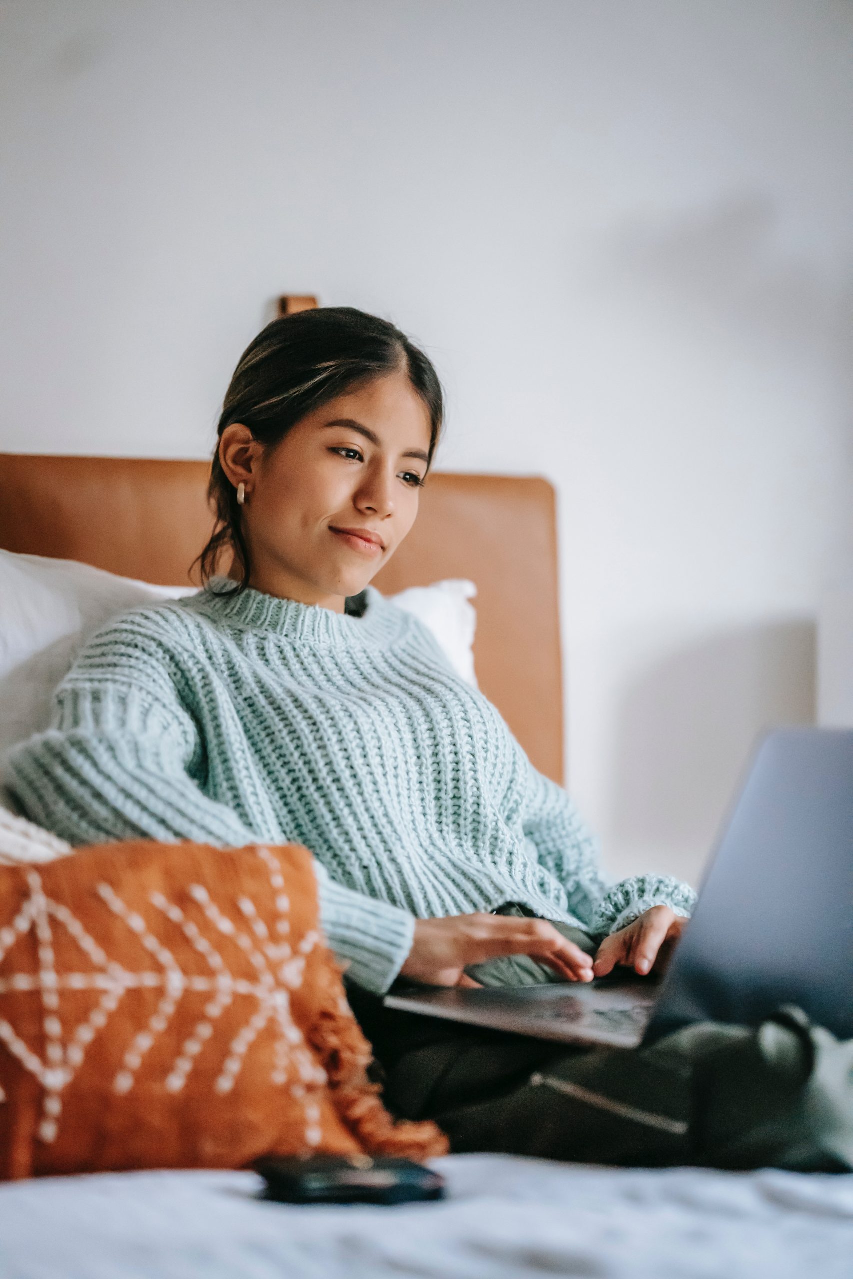 Millennial Latinx woman attends an online therapy session while sitting on her bed. She is holding a laptop on her lap and has an engaged, interested expression on her face.