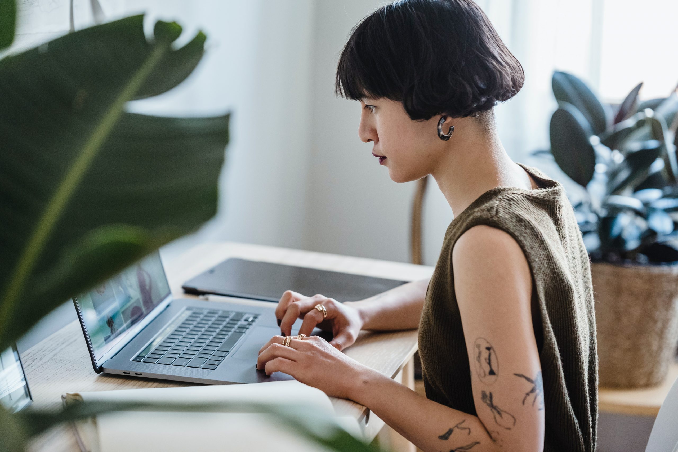 A queer nonbinary Asian person with short hair and tattoos sits in front of their laptop at a desk while working from home.