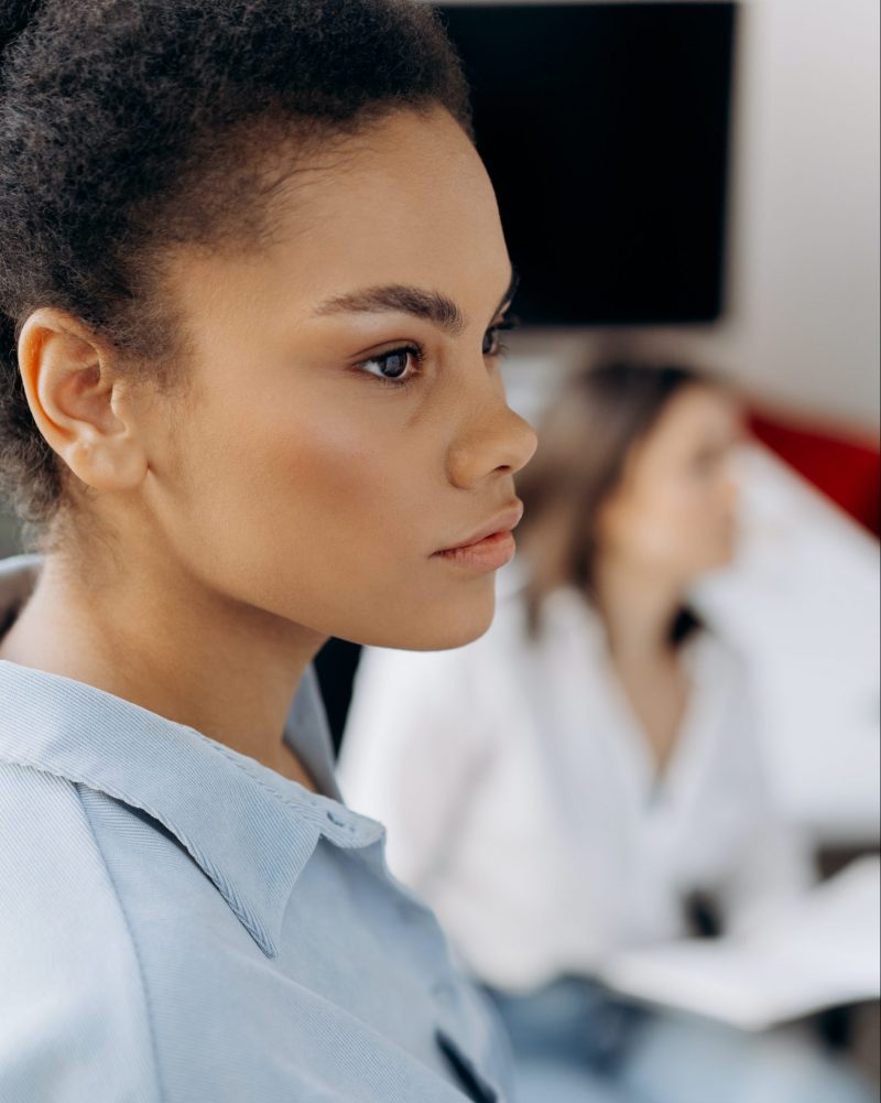 Young Black tech professional woman with natural hair and a light blue blouse, photographed in profile. She has a pensive expression on her face.