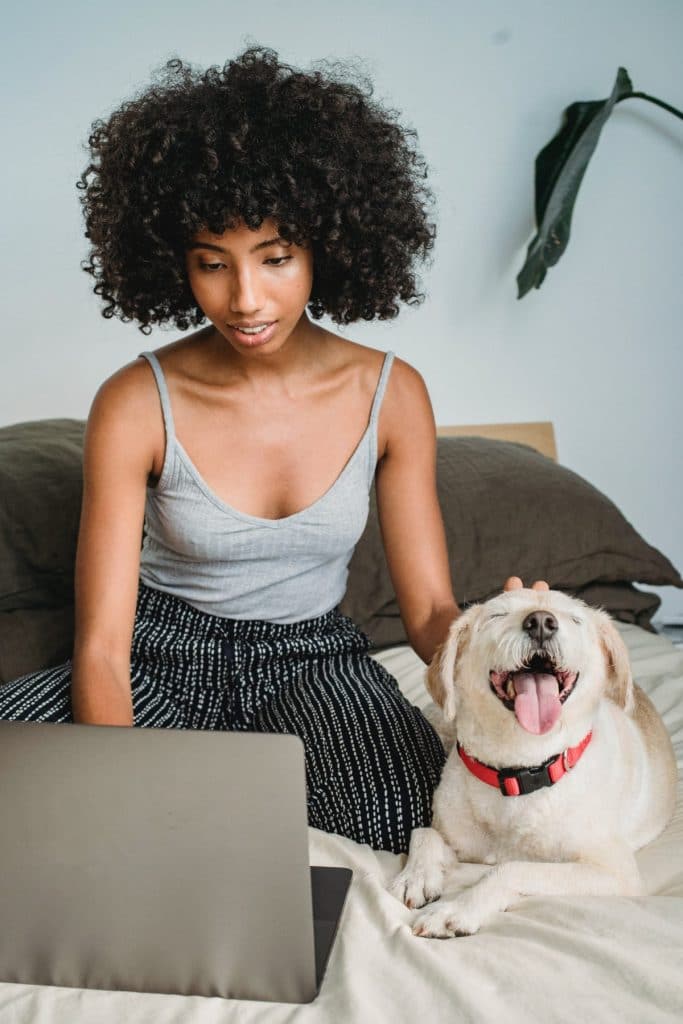 A young African American woman sits on her bed with a friendly dog. She is attending an online therapy session on her laptop.