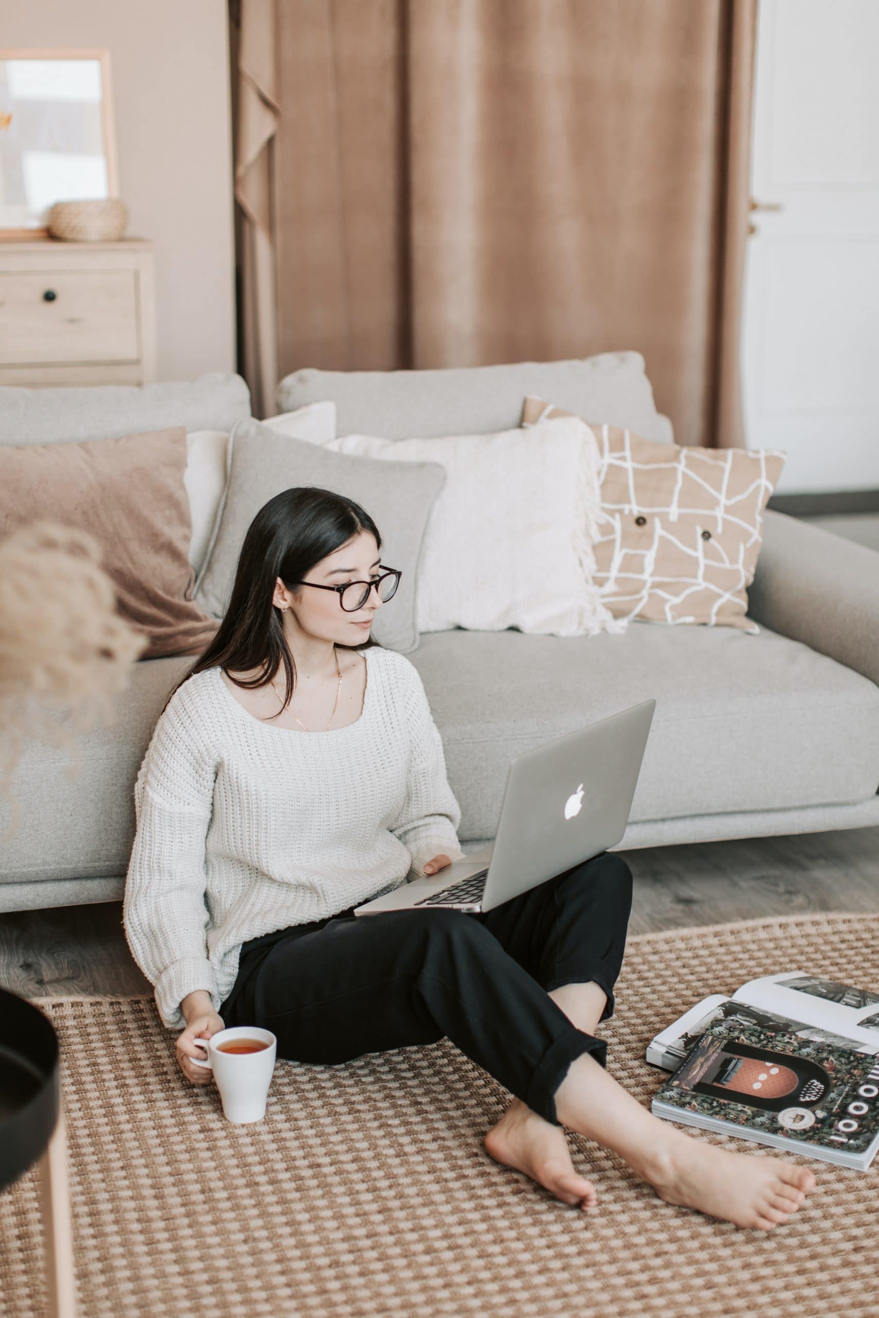 Young tech professional woman, sitting on the floor in front of her sofa. She is attending an online therapy session from home on her laptop. She has a serious look on her face and is holding a cup of tea.