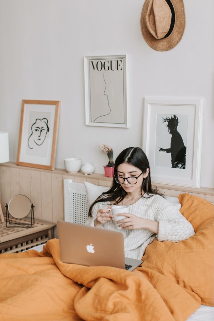 Woman with glasses and long black hair attends an online therapy session on her laptop from bed.