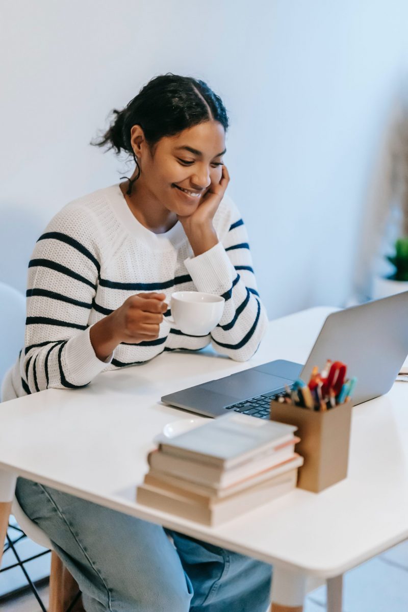 A South Asian millennial woman drinks a cup of coffee at her desk while working.
