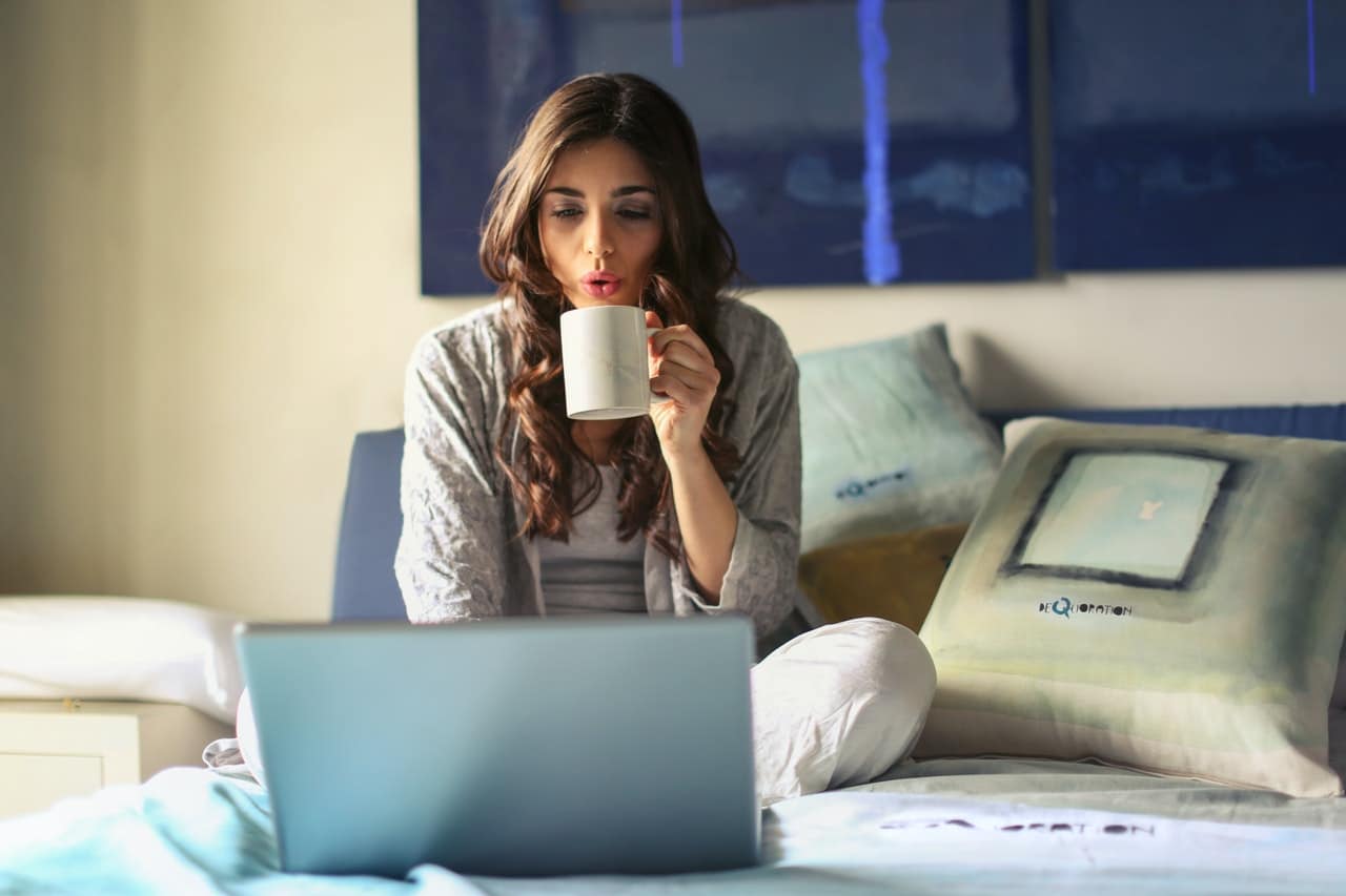 Woman attends an online therapy session at her laptop. She is sitting on top of her bed and blowing on a cup of coffee, as though to cool it off.