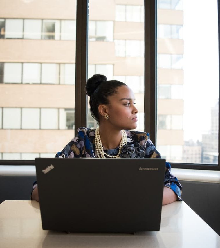 Black woman in professional attire looks out her office window with a serious expression on her face.