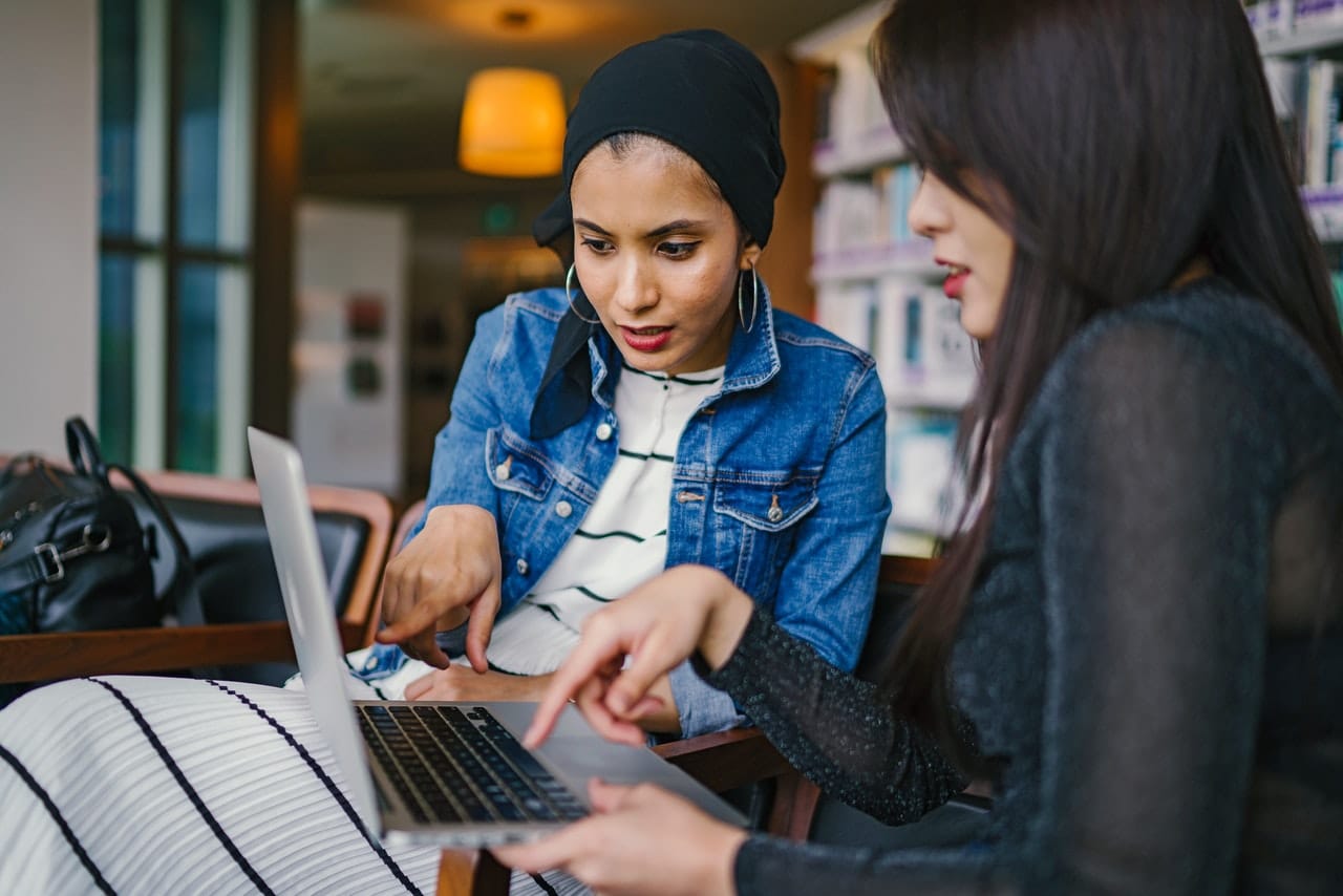 Two Millennial women looking at a laptop together. The first is a brown skinned women with a head scarf and hoop earrings. The second is an Asian American woman with long hair and red lipstick.