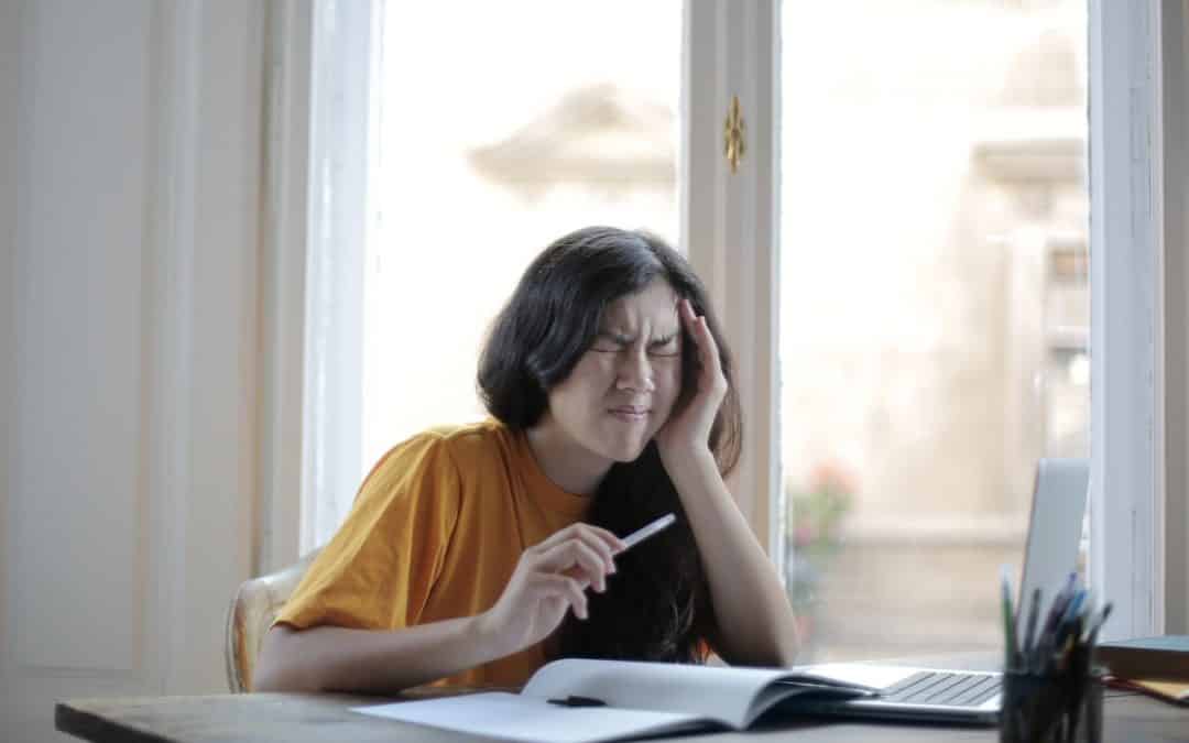 Woman with headache working at computer with a book open in front of her. She appears to be an Asian American millennial woman with long hair and glasses.