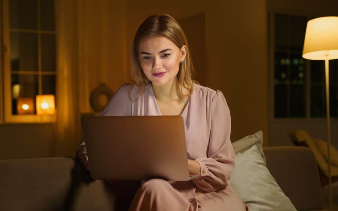 A woman in a mauve dress sits in a dimly lit room on her laptop, attending an online therapy session.