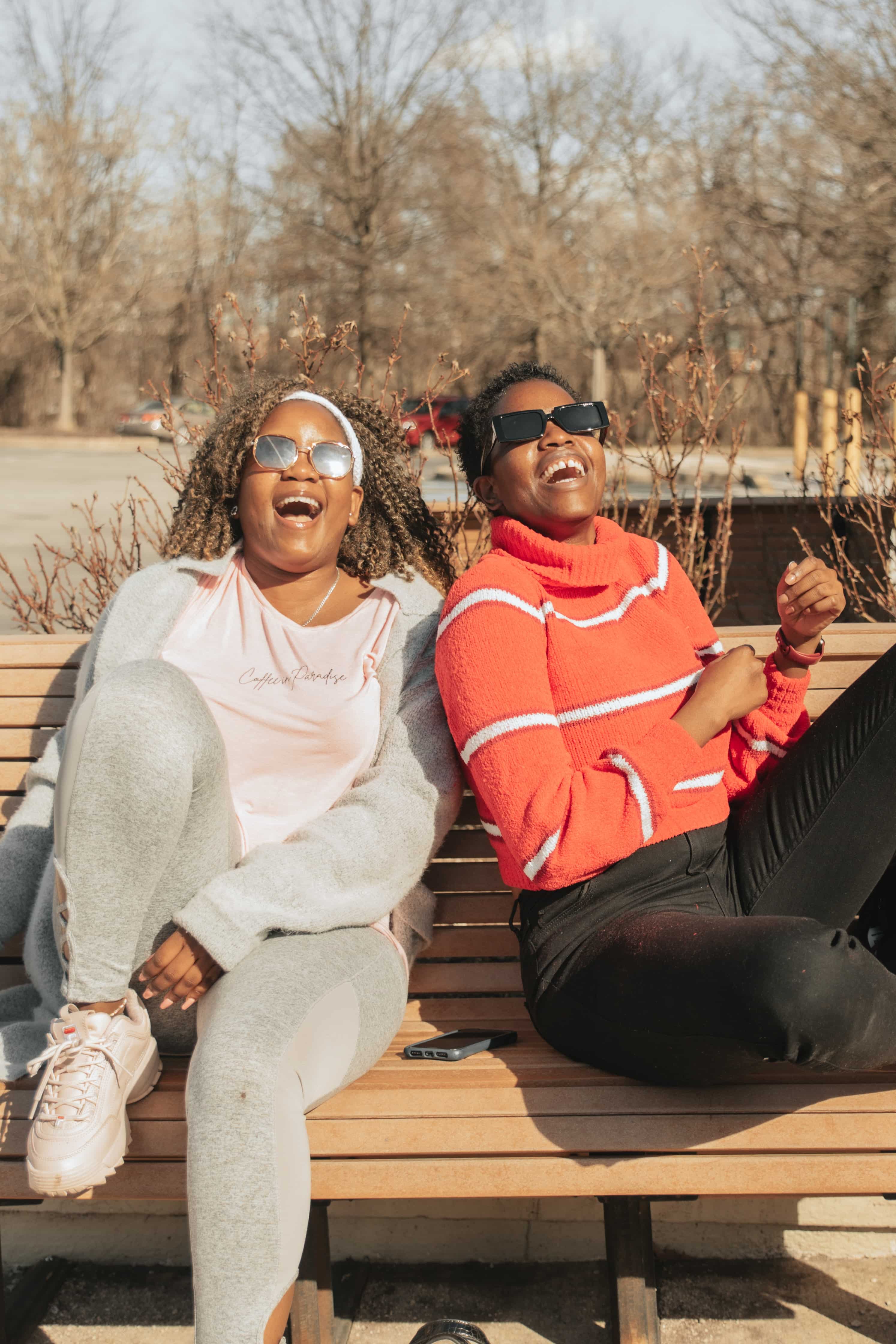 Two friends sit together on a park bench on a winter day. They are both Black women, wearing sunglasses and casual, warm clothing. They both have their heads tilted back and are laughing