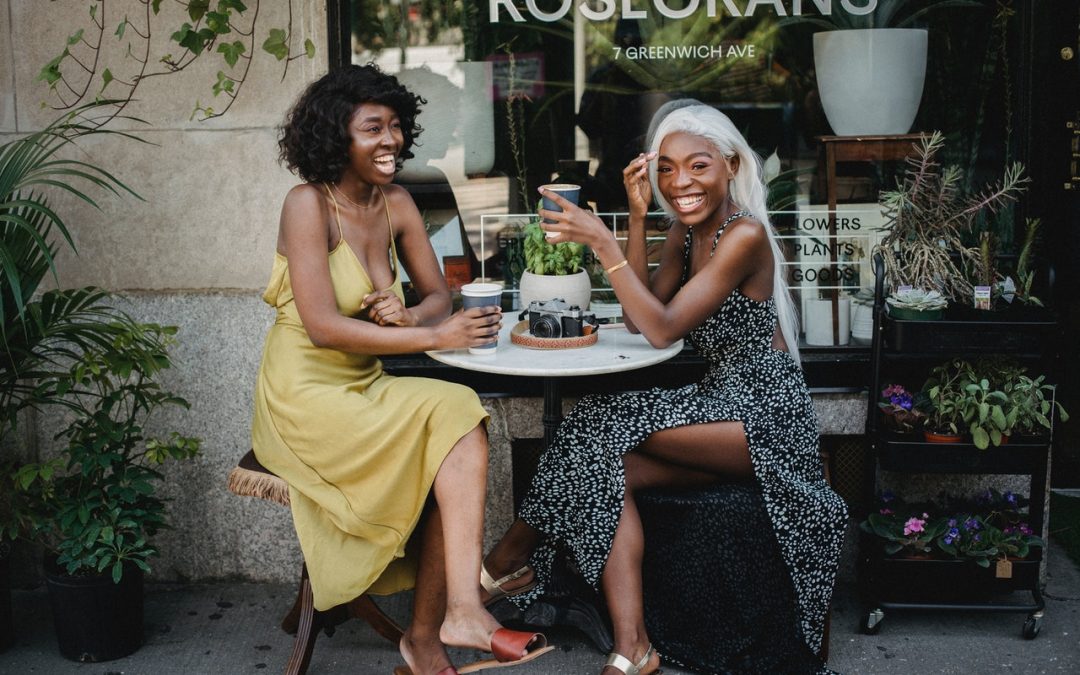 Two young Black women sit at a cafe table. They are both wearing sundresses and have a happy expression on their faces.