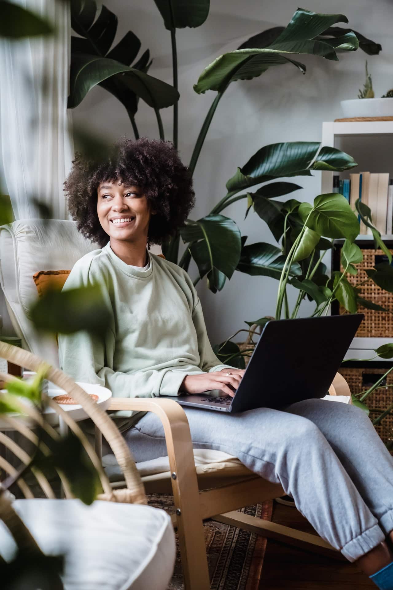 A young Black woman in casual clothing sits in an armchair with a laptop on her lap. She has a relaxed expression on her face. The room is full of houseplants and natural lighting.