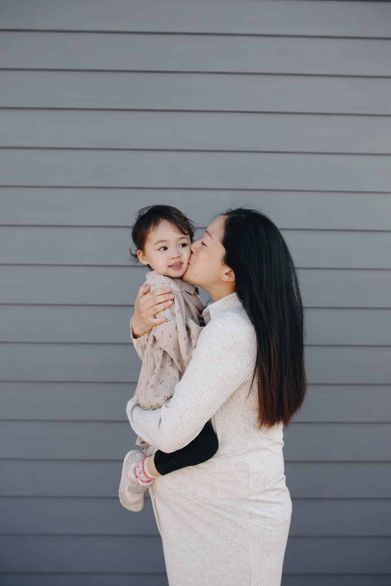 Pregnant Asian American mom holding and kissing a baby outdoors in front of a gray wall.