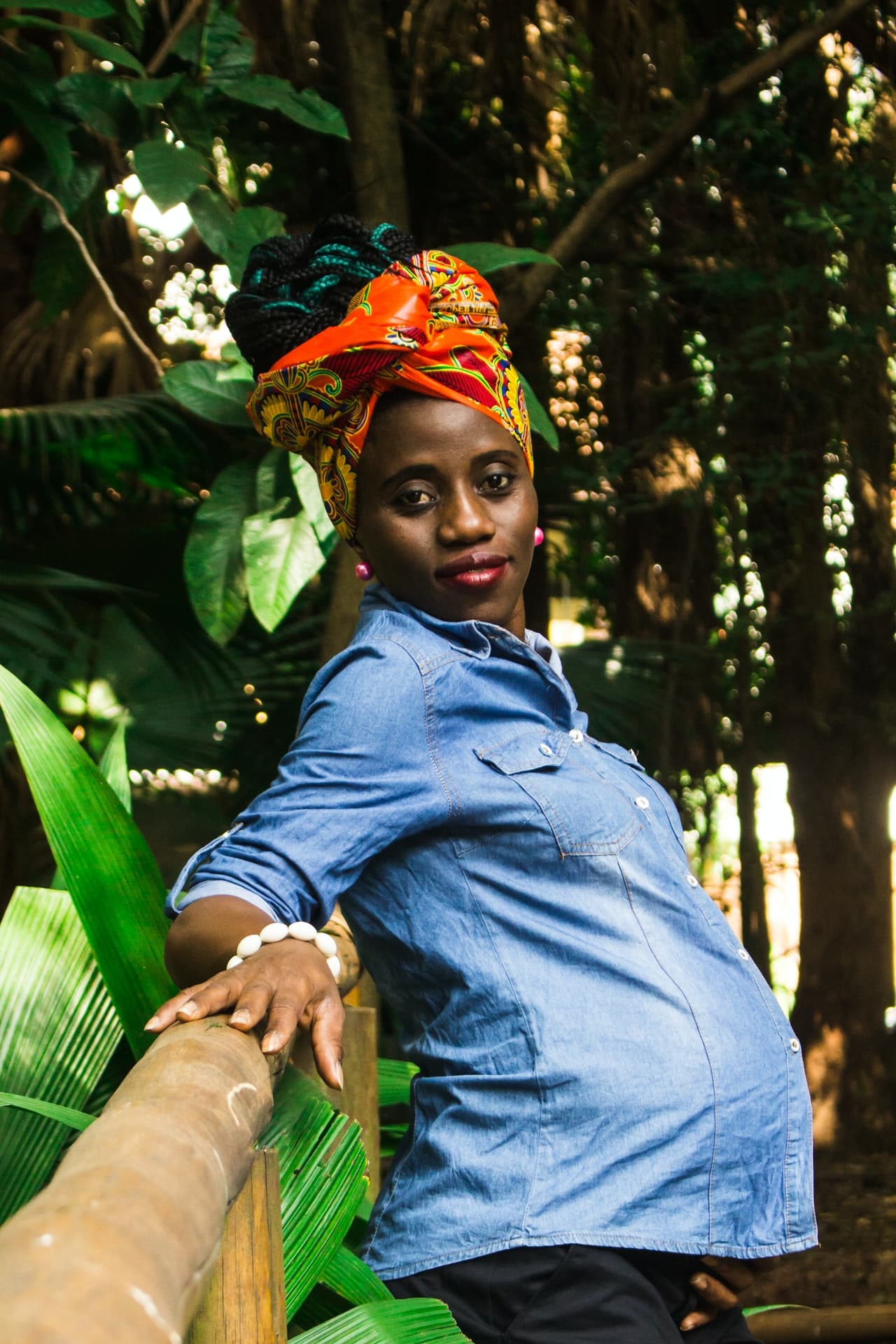 Pregnant woman poses in front of a fence outdoors. She is a Black woman in a blue button down and a red head wrap.