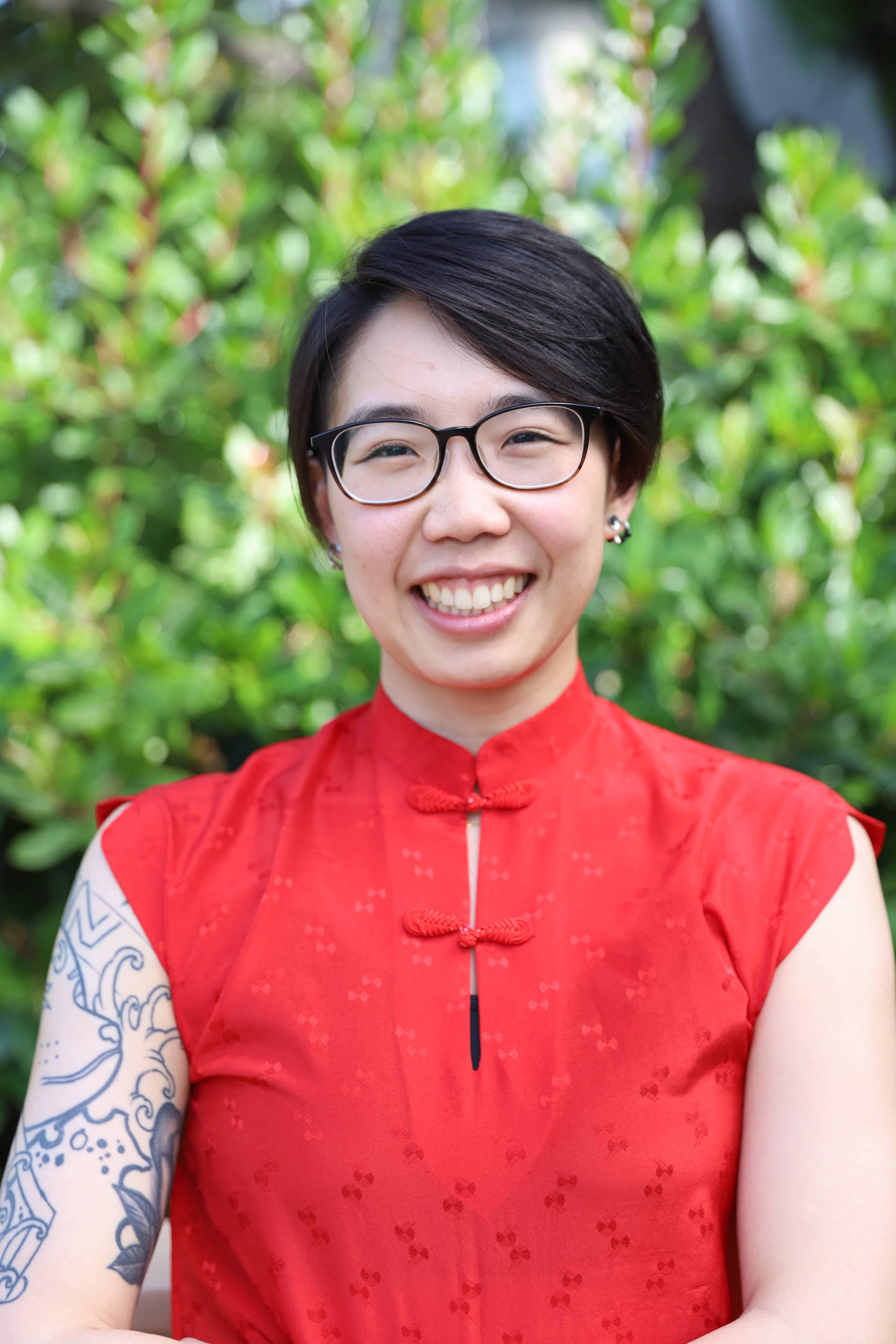 Headshot of Dr. Justine Fan (ze/zer), an Asian American psychologist with short hair in a red blouse. A half sleeve tattoo is visible.