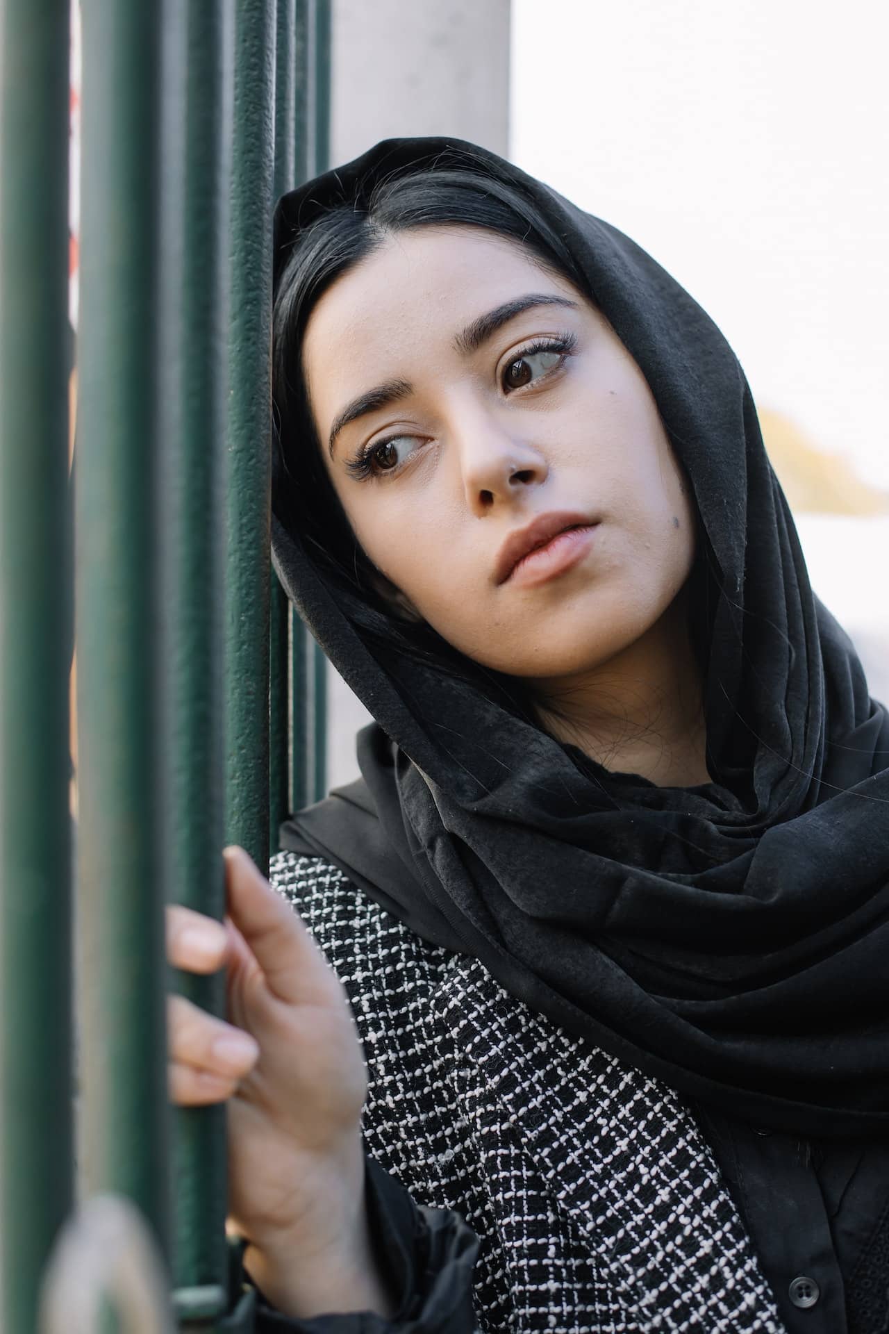 A woman in a dark coat and black hijab rests her head against a gate with a melancholy expression.
