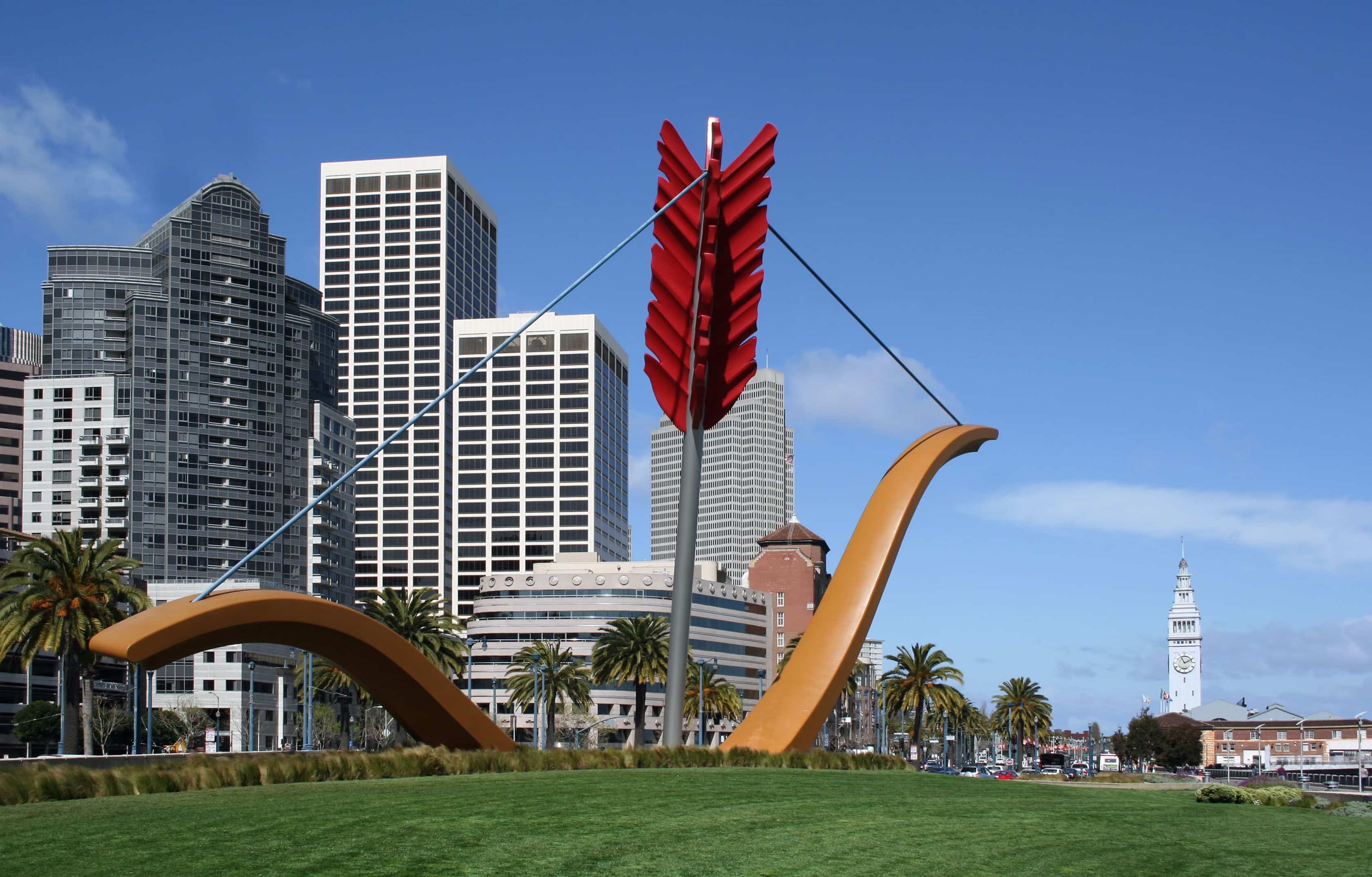 A photo of Cupid's Bow sculpture, a giant bow and arrow on the Embarcadero in San Francisco. The ferry building and downtown can be seen in the background.