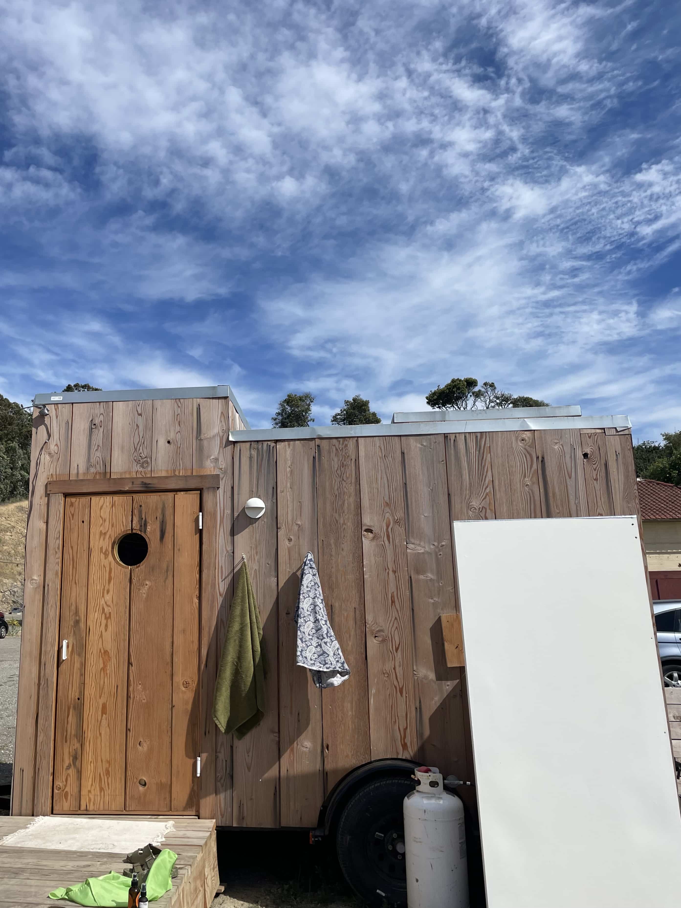 Photo of a small wooden structure with towels hanging from hooks, shot against a blue sky.