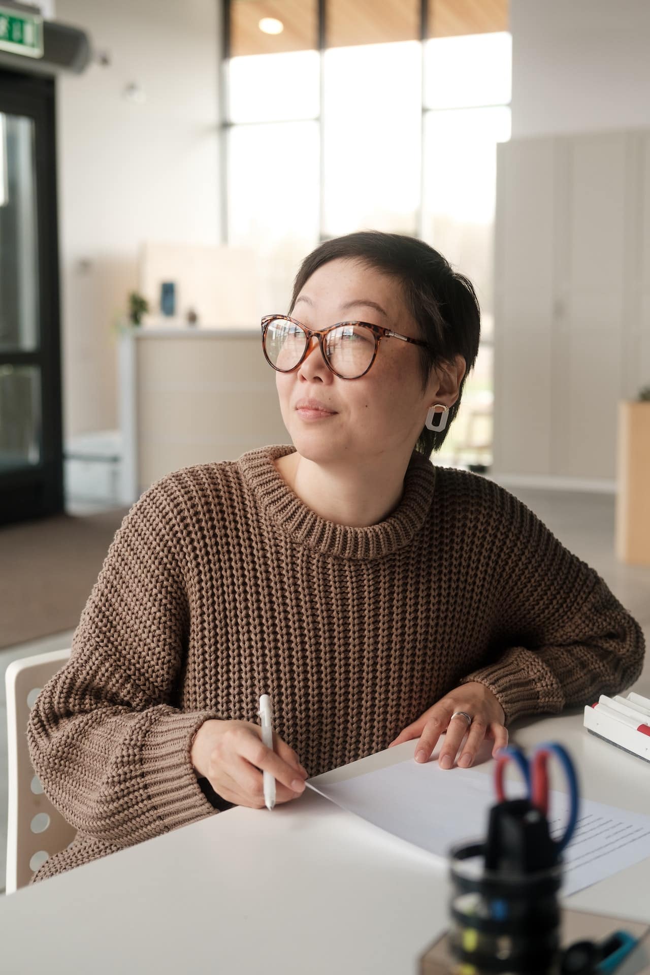 Asian American person with short hair and glasses looking up reflectively while writing