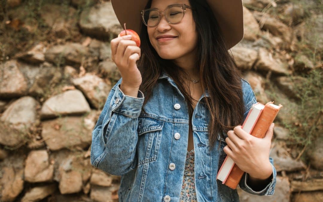 Happy looking Asian woman with long hair and glasses eating an apple and carrying a book.