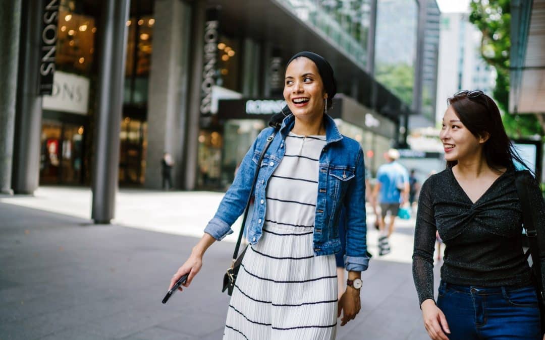 Two women walking together in a city in casual attire, with happy expressions on their face.
