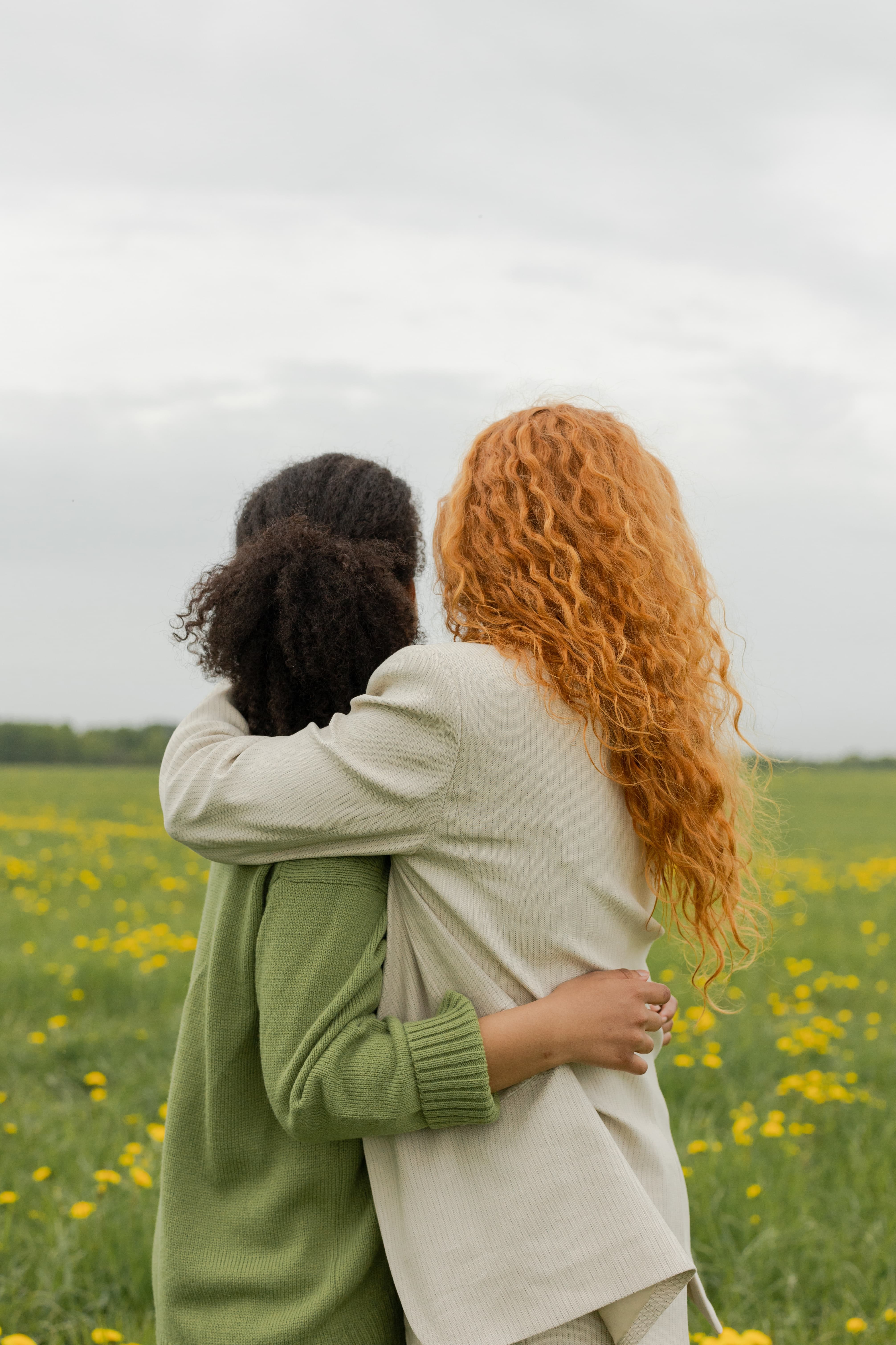 Two friends standing with their arms around each other, photographed from behind. One has bright red hair, the other has natural black hair.