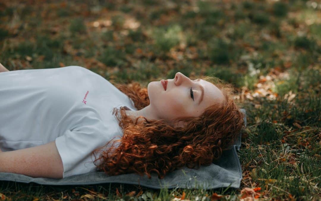 A woman with red hair lies on a yoga mat outdoors in the shade.