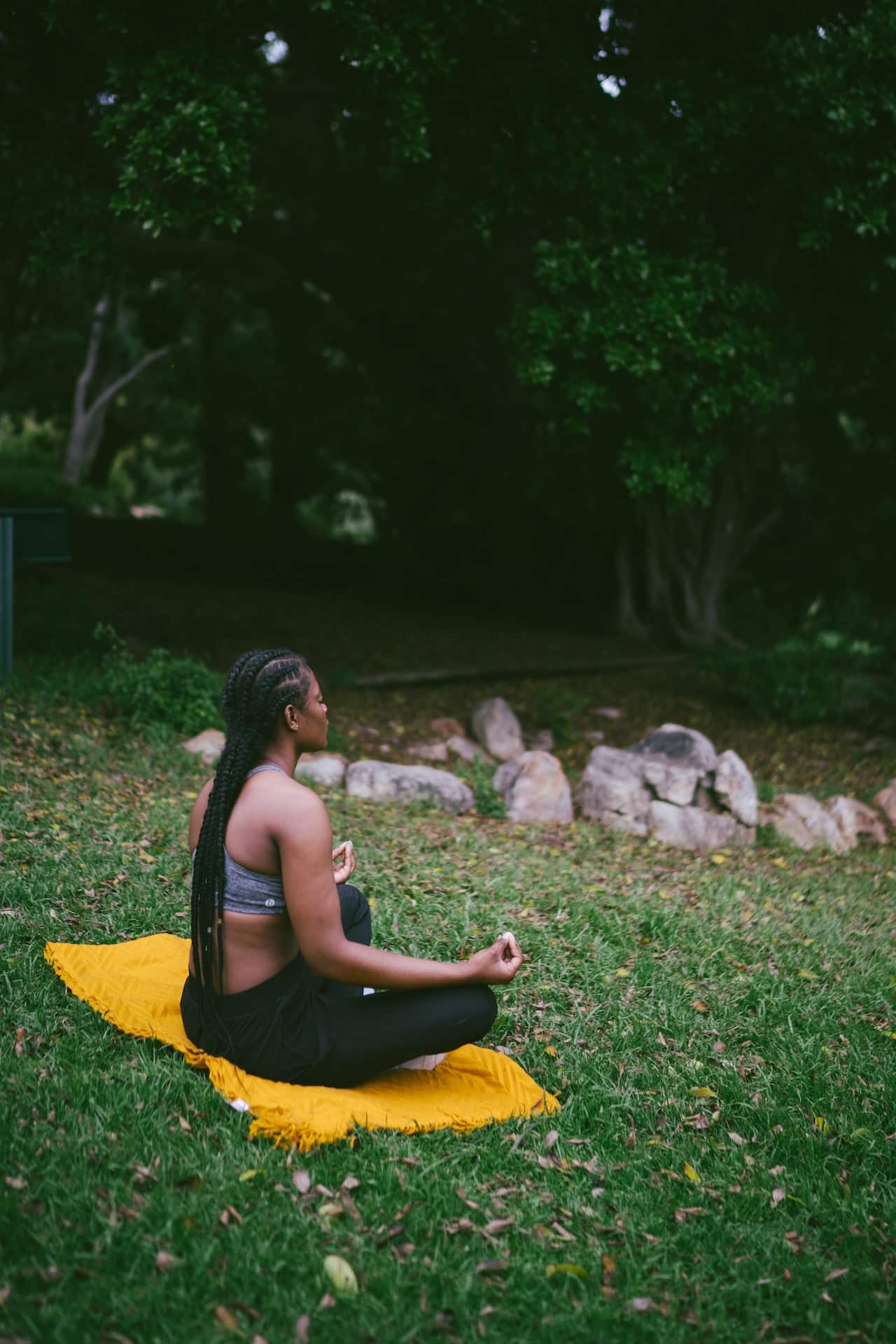 A black woman with long braids is sitting cross-legged on a blanket meditating outdoors in sweatpants and a sports bra.