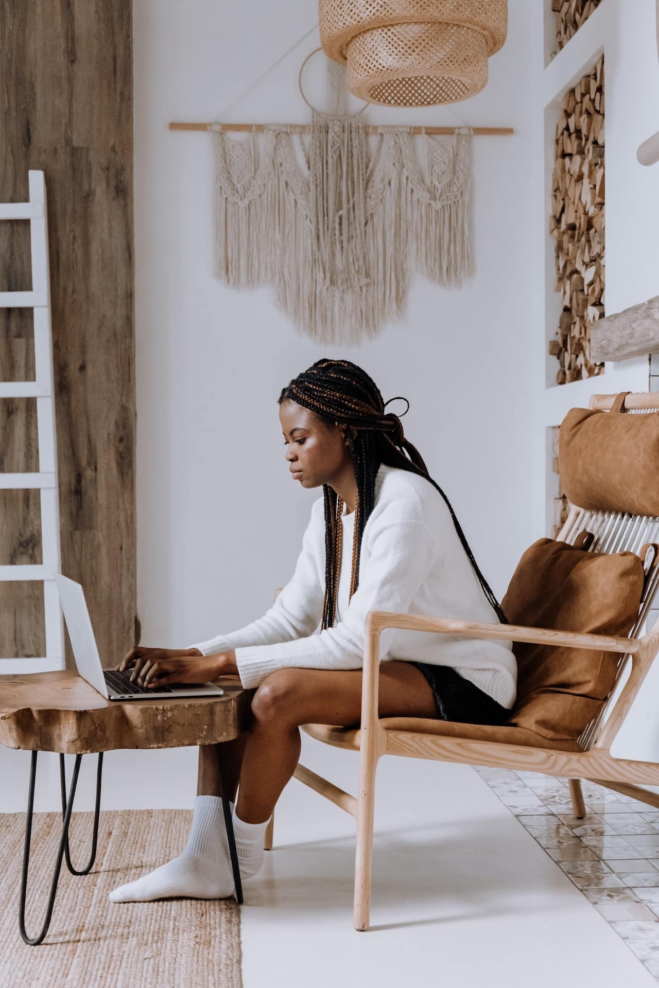 A Black femme presenting person sits at their laptop, apparently attending an online therapy session from home in a tastefully furnished living room. They have long braids and are wearing a white sweater, shorts and socks.n 