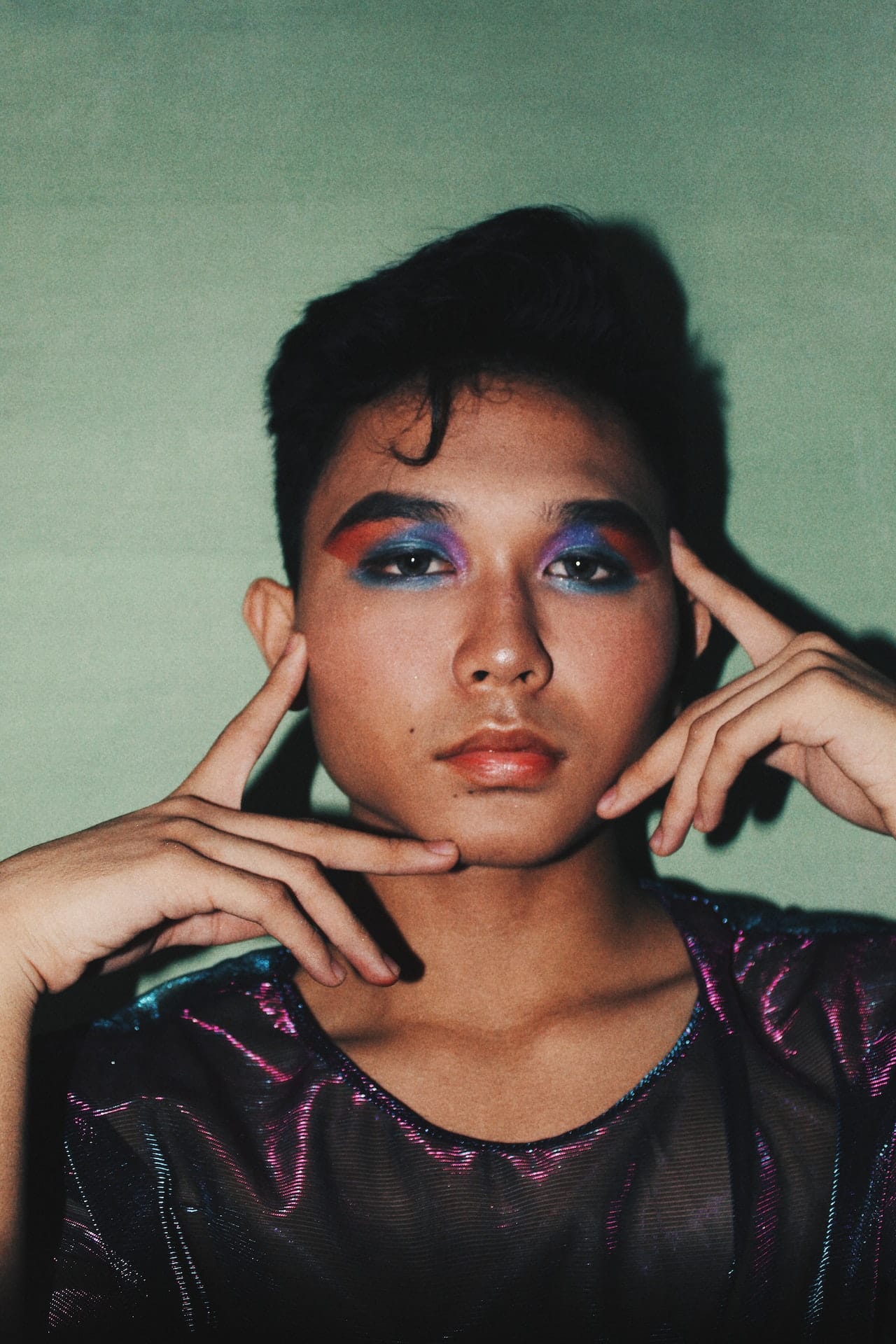 A close up photo of a young queer person with brown skin and dark short hair. They are wearing brightly colored eye makeup and framing their face with their hands.