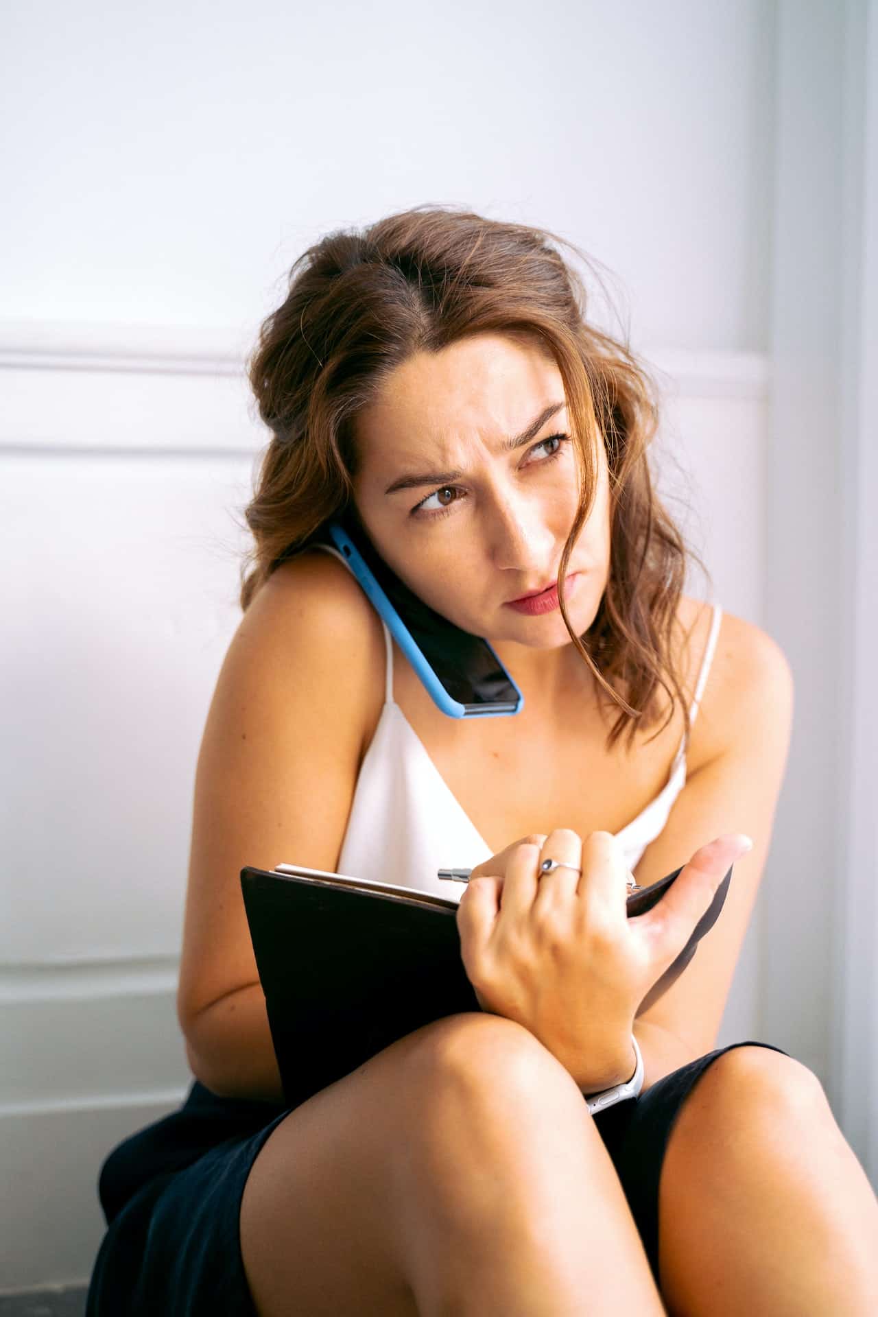 A woman sits on the floor against the wall with a frustrated look on her face. She is on a cell phone and has a notebook in her lap.