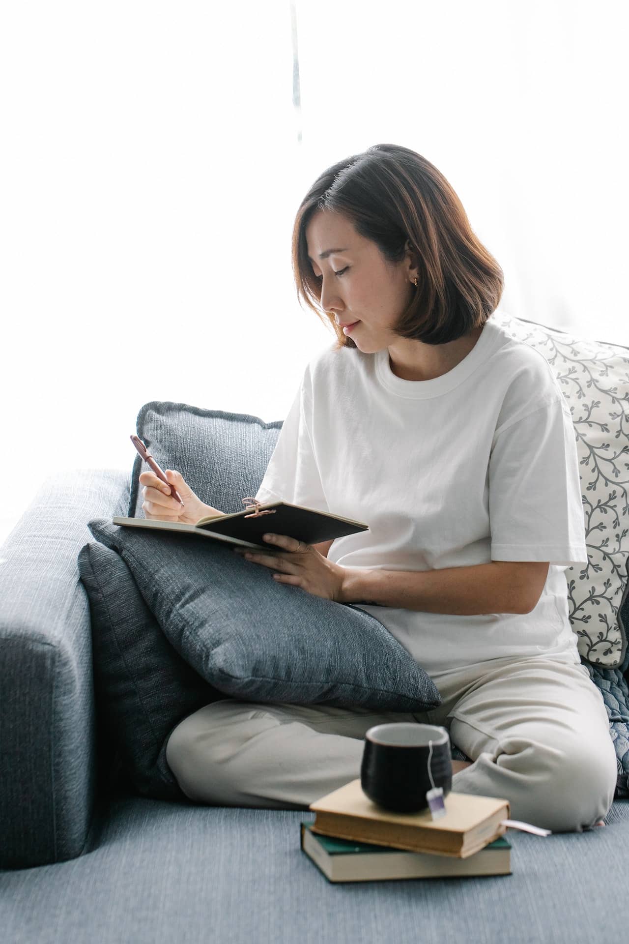Asian American woman sits on a sofa by the window writing in her jorunal. A cup of tea and some books are in the foreground.