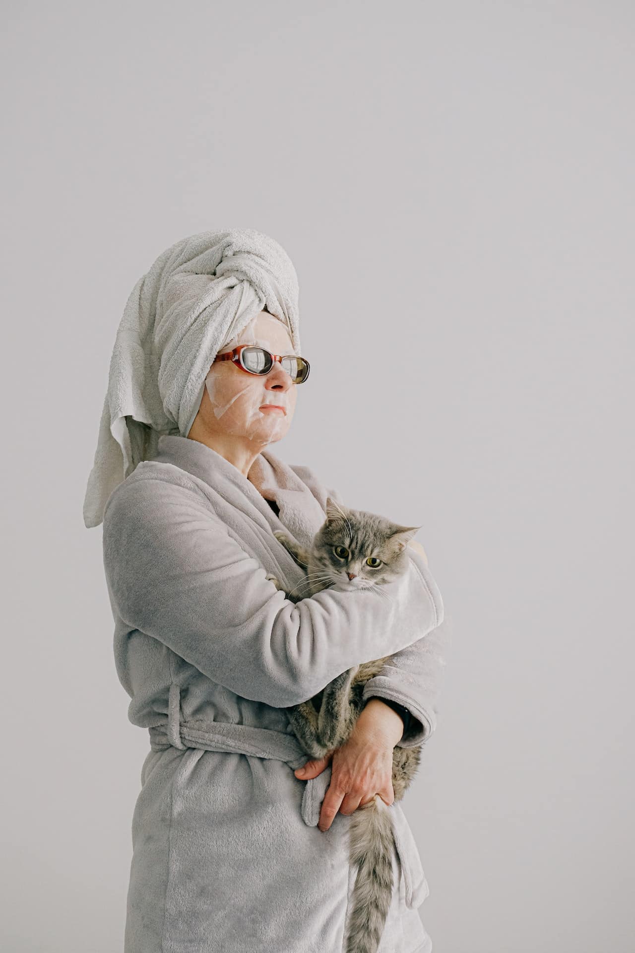 A woman in a robe holds a cat. She is wearing a hydrating face mask and has a towel wrapped around her hair.