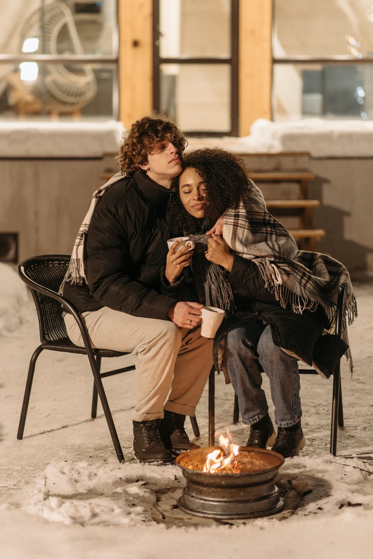 A young couple cuddles outdoors by a fire, wrapped in blankets.