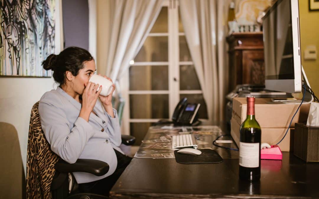 A woman sits at a desk in a blazer, drinking out of a mug while looking towards her computer screen. A bottle of wine is seen in the foreground.
