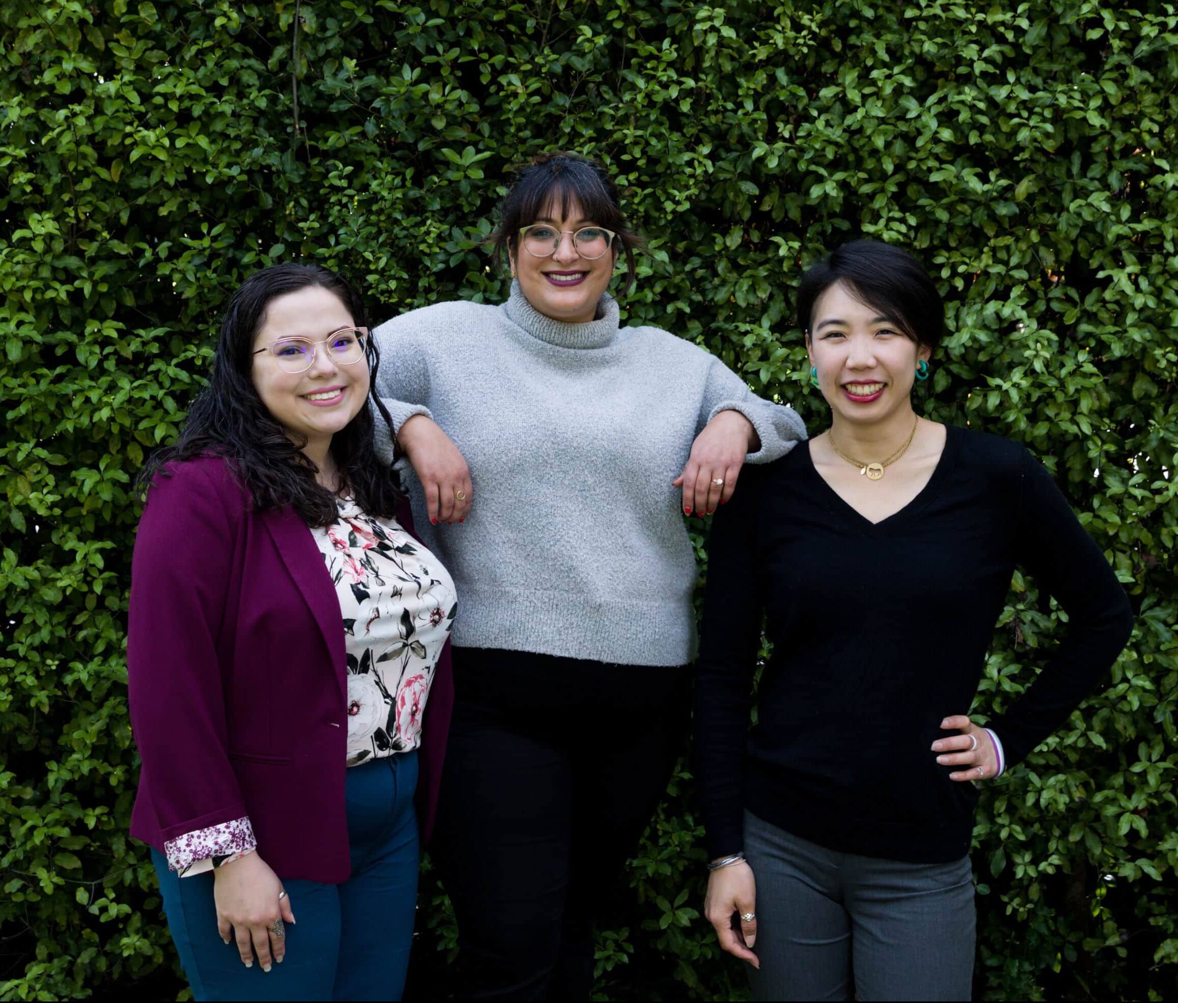 A group of three Stella Nova San Francisco therapists pose with smiles in front of a green hedge.