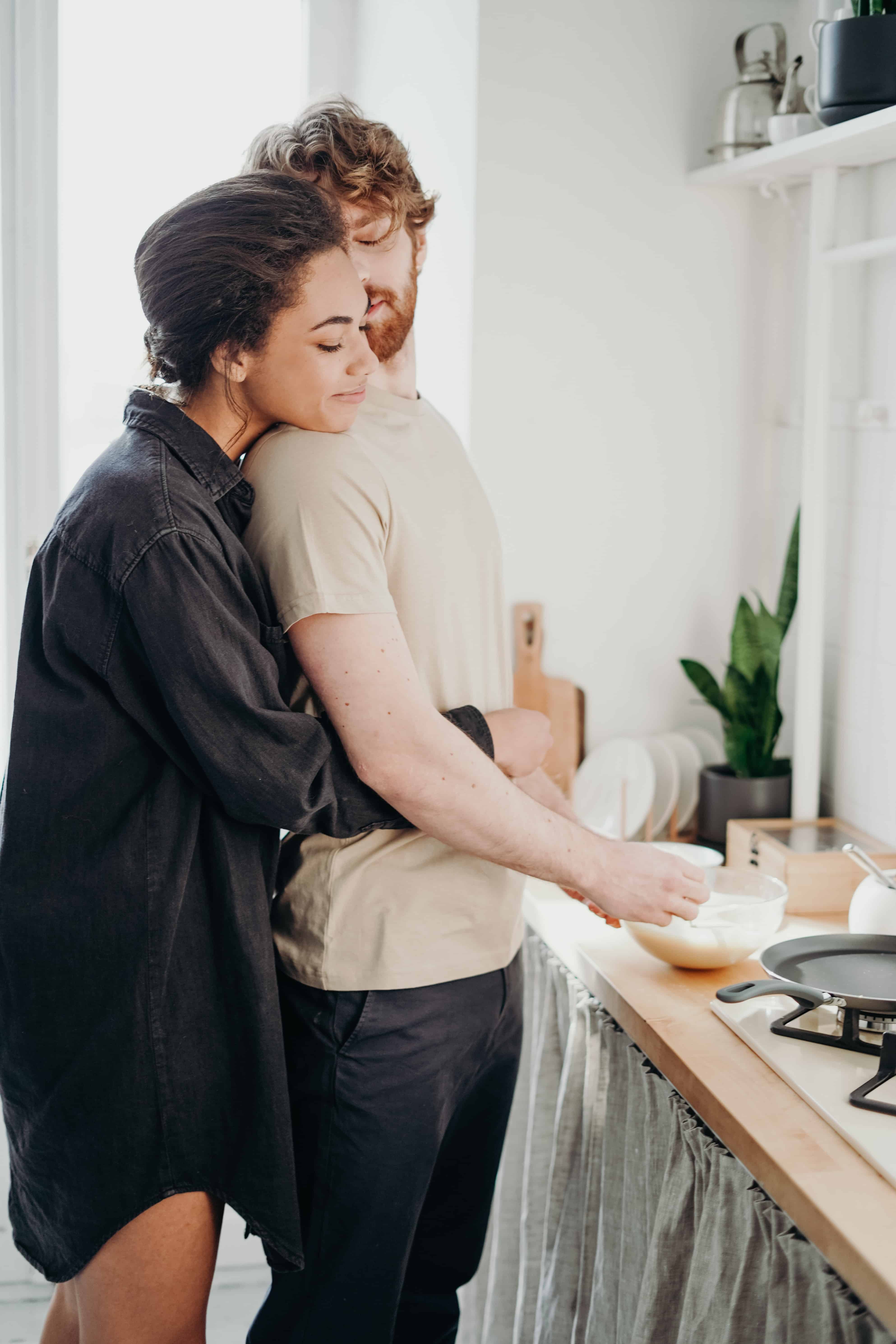 Photo of a couple making cooking together in their kitchen. One partner is preparing food while the other embraces him from behind.