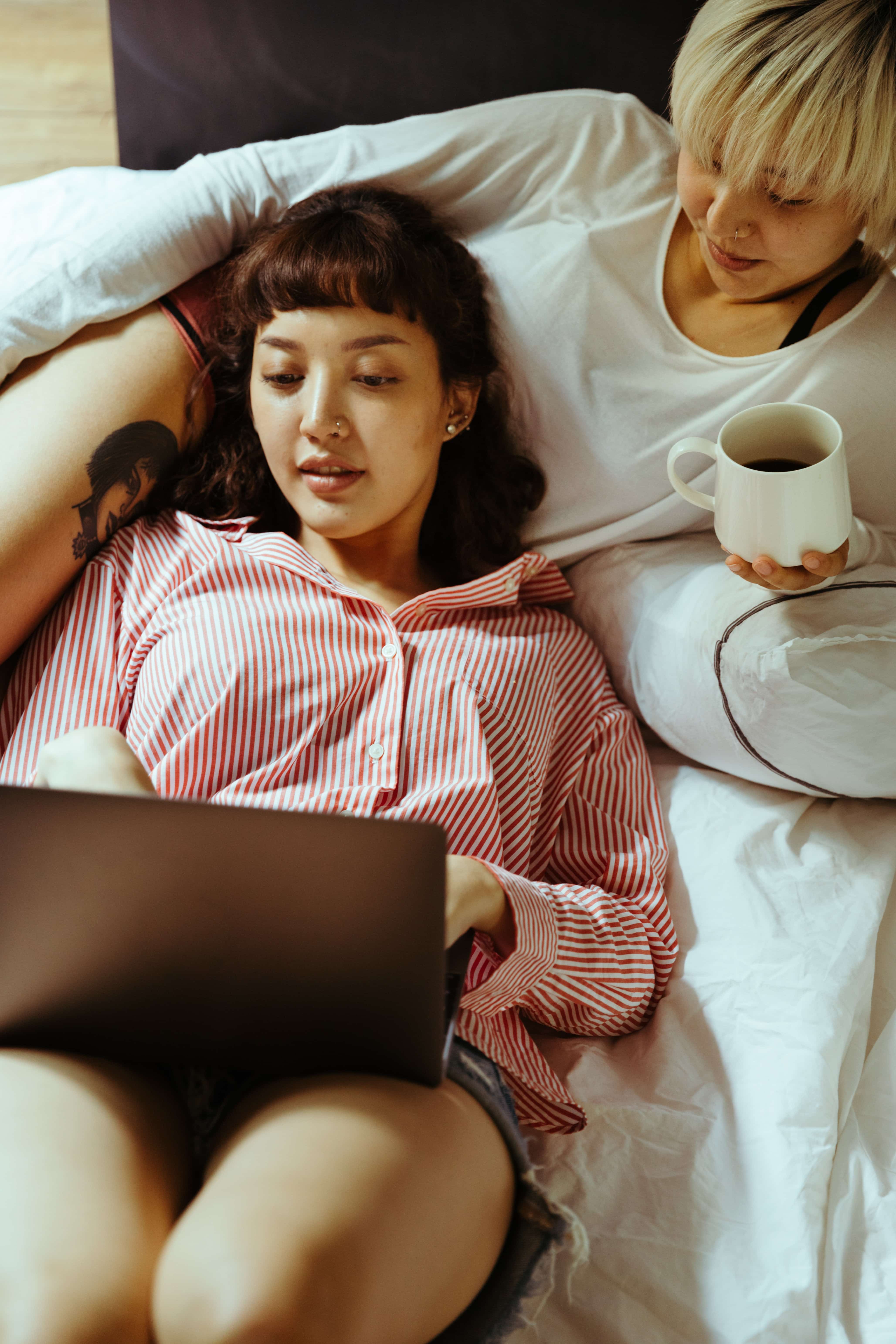 A queer couple relaxes in bed together. One is drinking a cup of coffee, while the other is using their laptop.