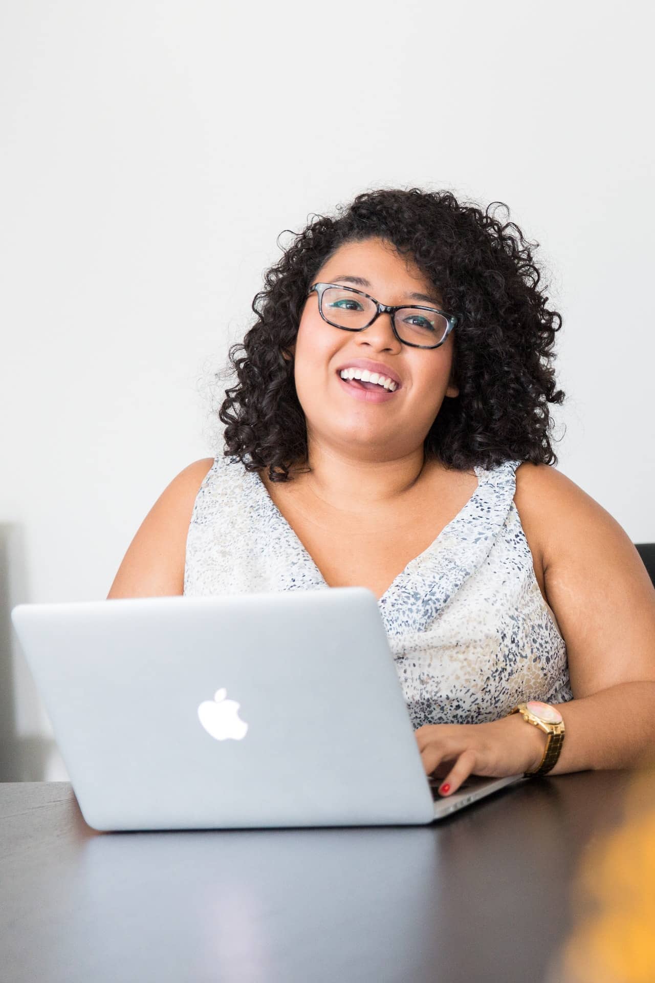 A curly haired woman with a medium complexion is sitting at her laptop smiling. She is wearing a sleeveless blouse and glasses. 