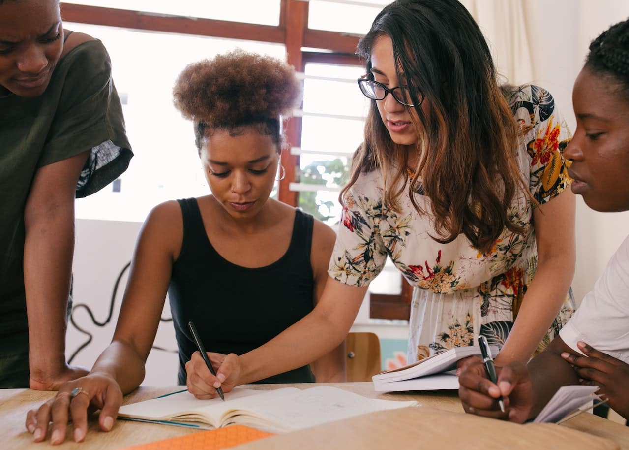 A group of women working together at a table. One is standing over the other, writing something in front of her.