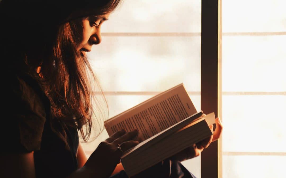 A woman sits at a window reading a book in soft lighting.