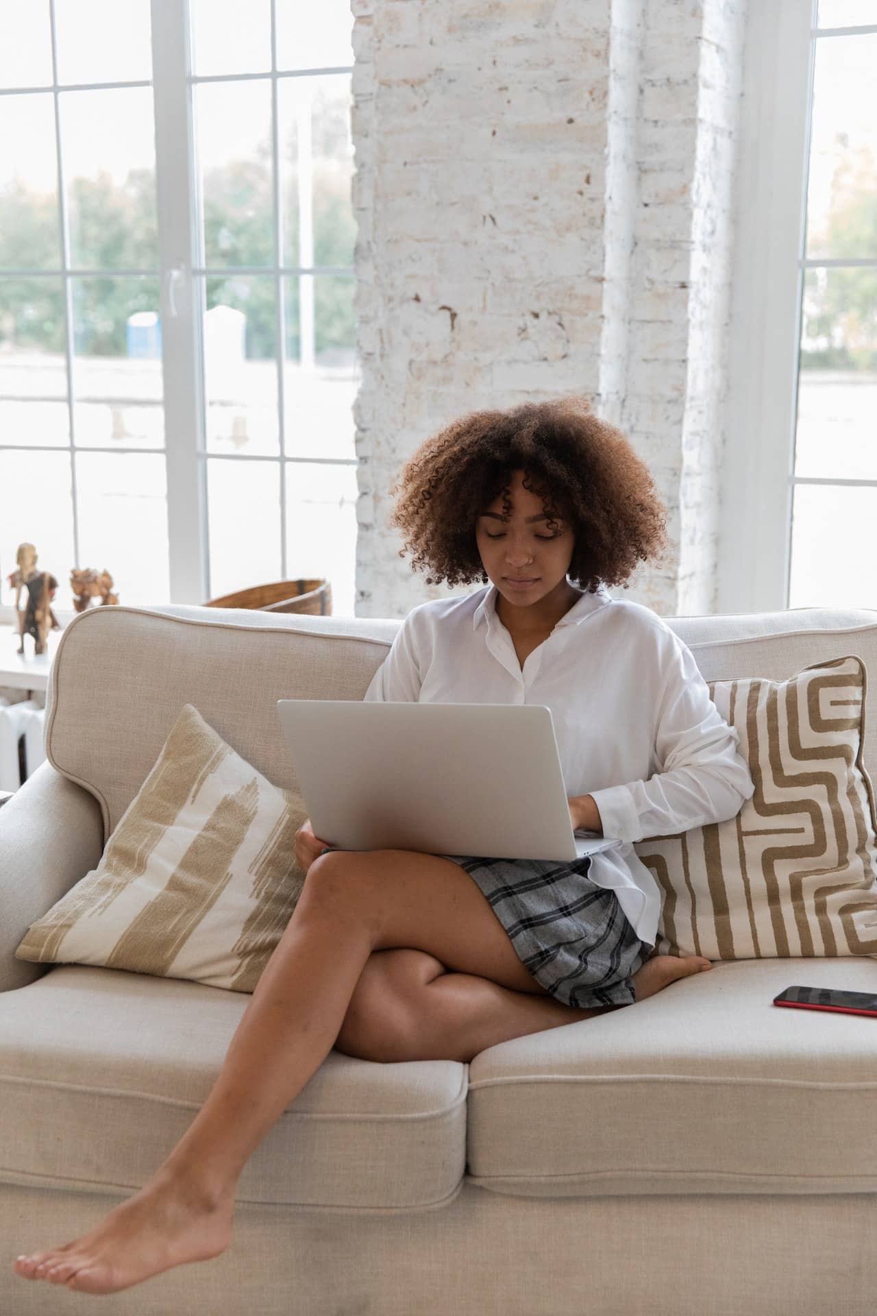 Black woman with chin length natural hair sits on her sofa in a white blouse and a casual skirt. She is working from home on her laptop.